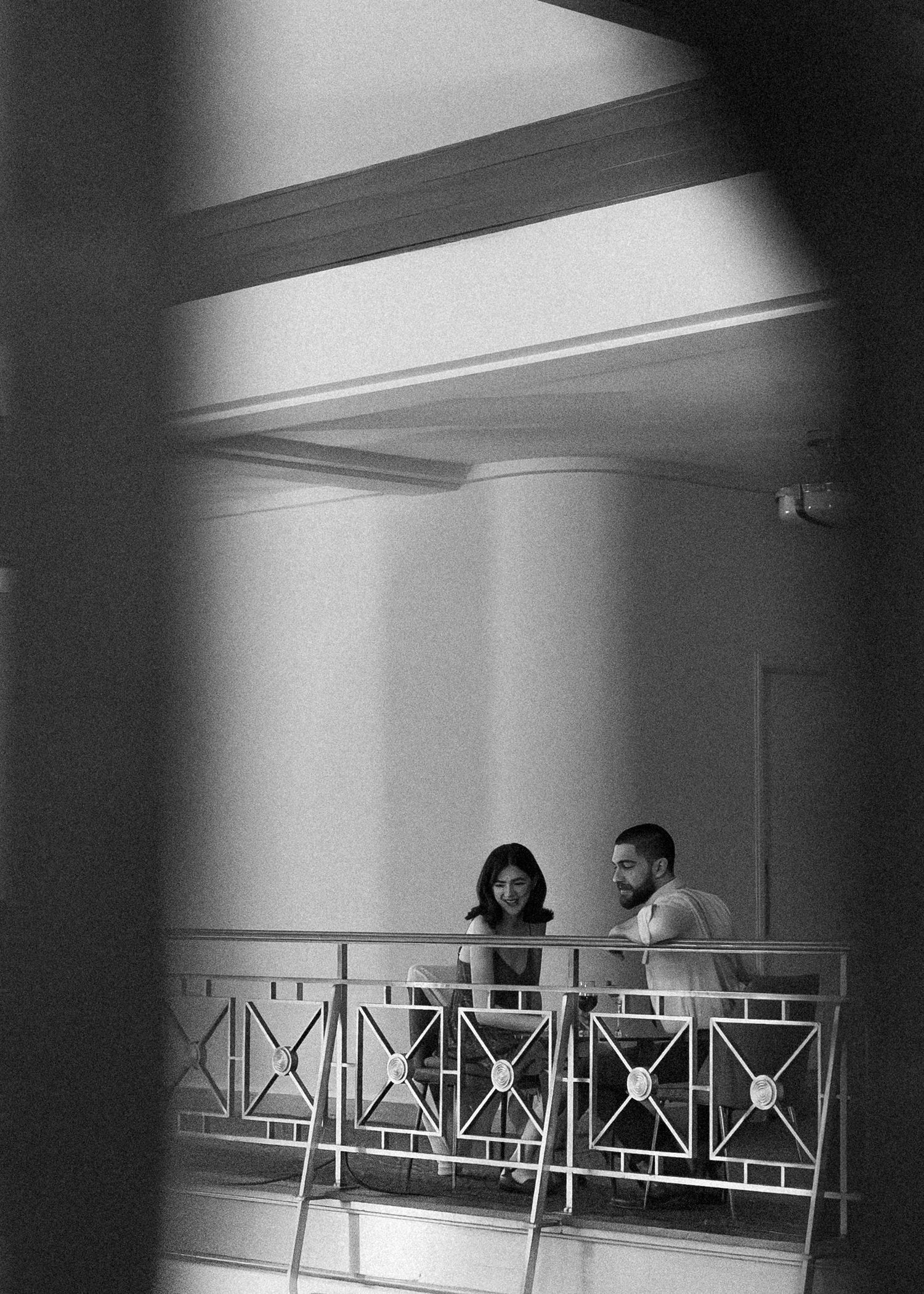 Black and white photo of a couple conversing on an ornate balcony railing, framed by dramatic shadows.