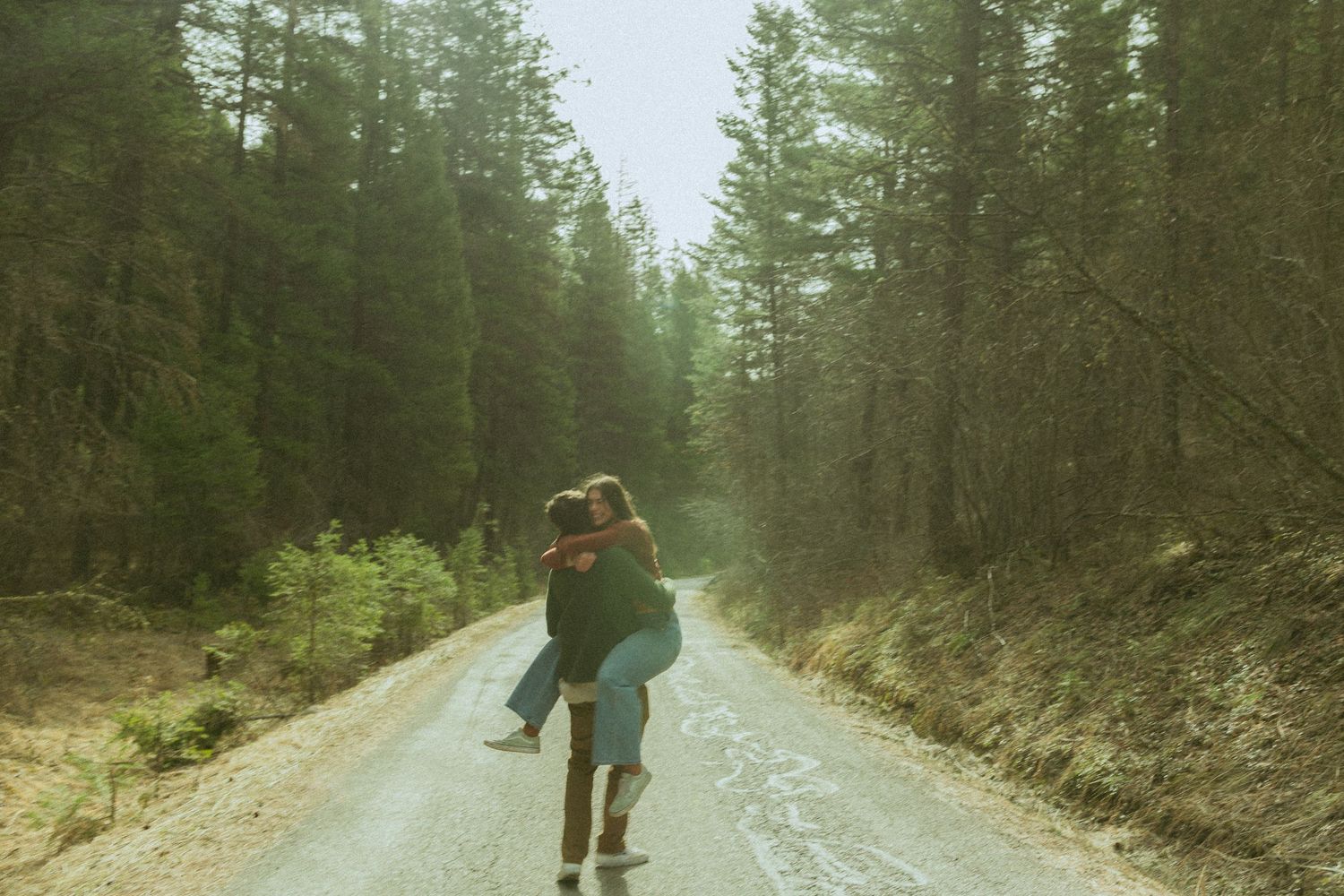 Couple sharing a romantic piggyback ride on a misty forest road surrounded by tall pine trees.