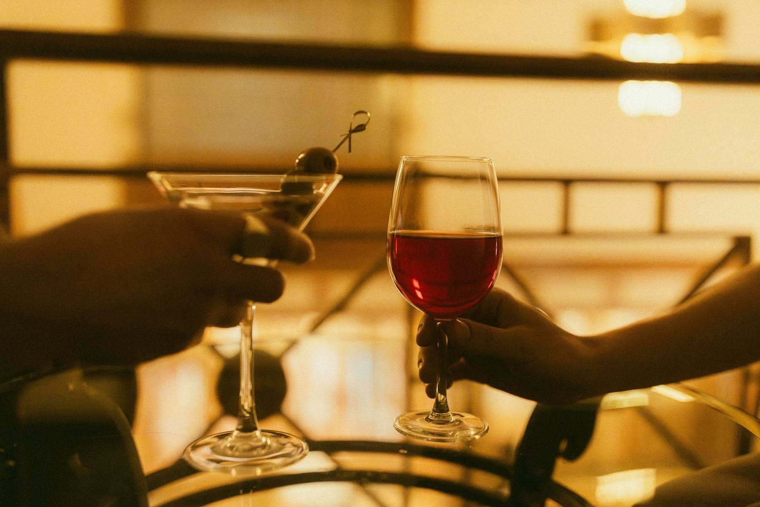 Two hands clinking glasses, one martini with olives and one red wine, in a warm golden-lit bar setting.