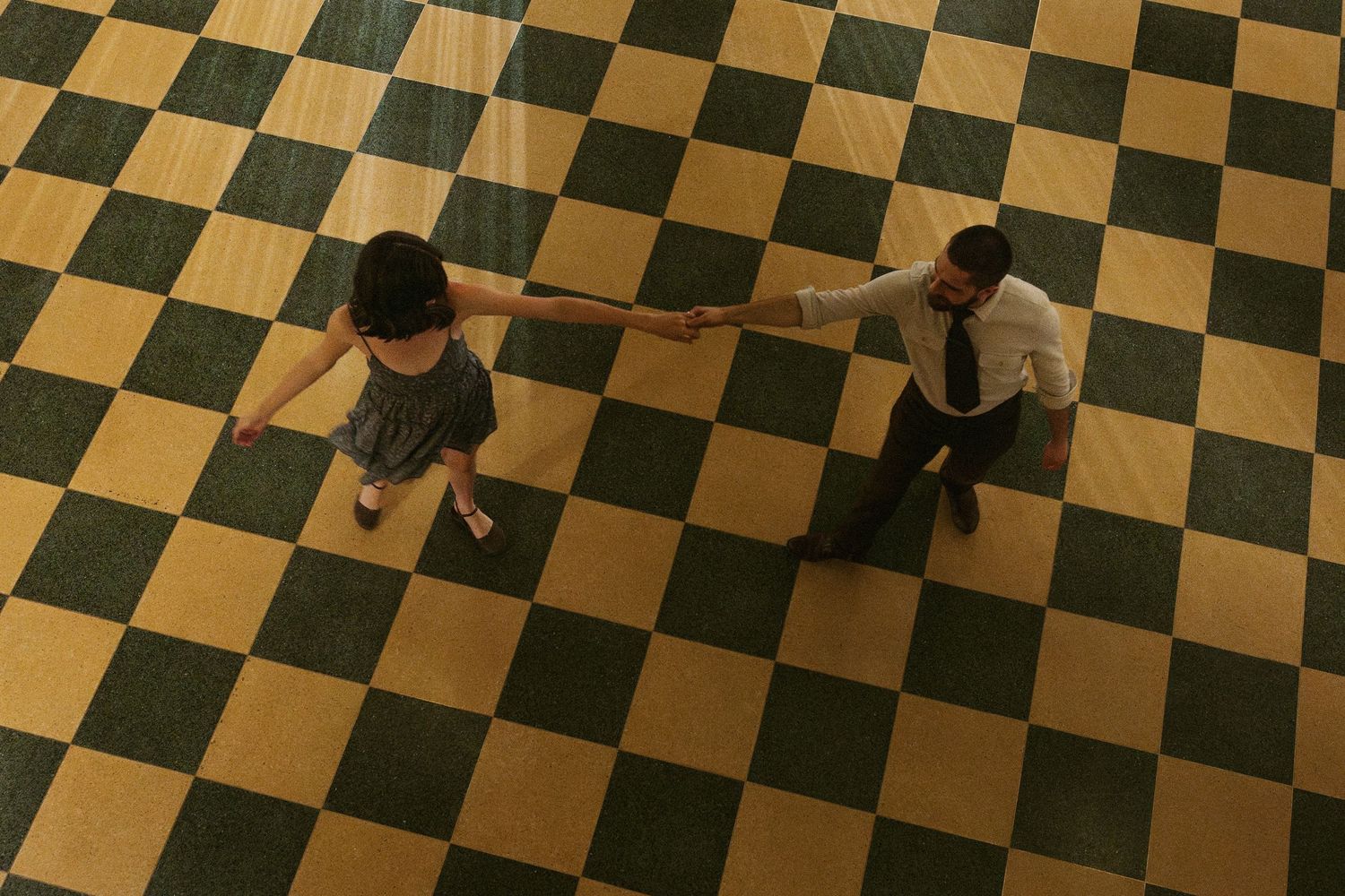 Couple dancing on a checkered green and gold floor, viewed from above, holding hands in elegant dance pose.