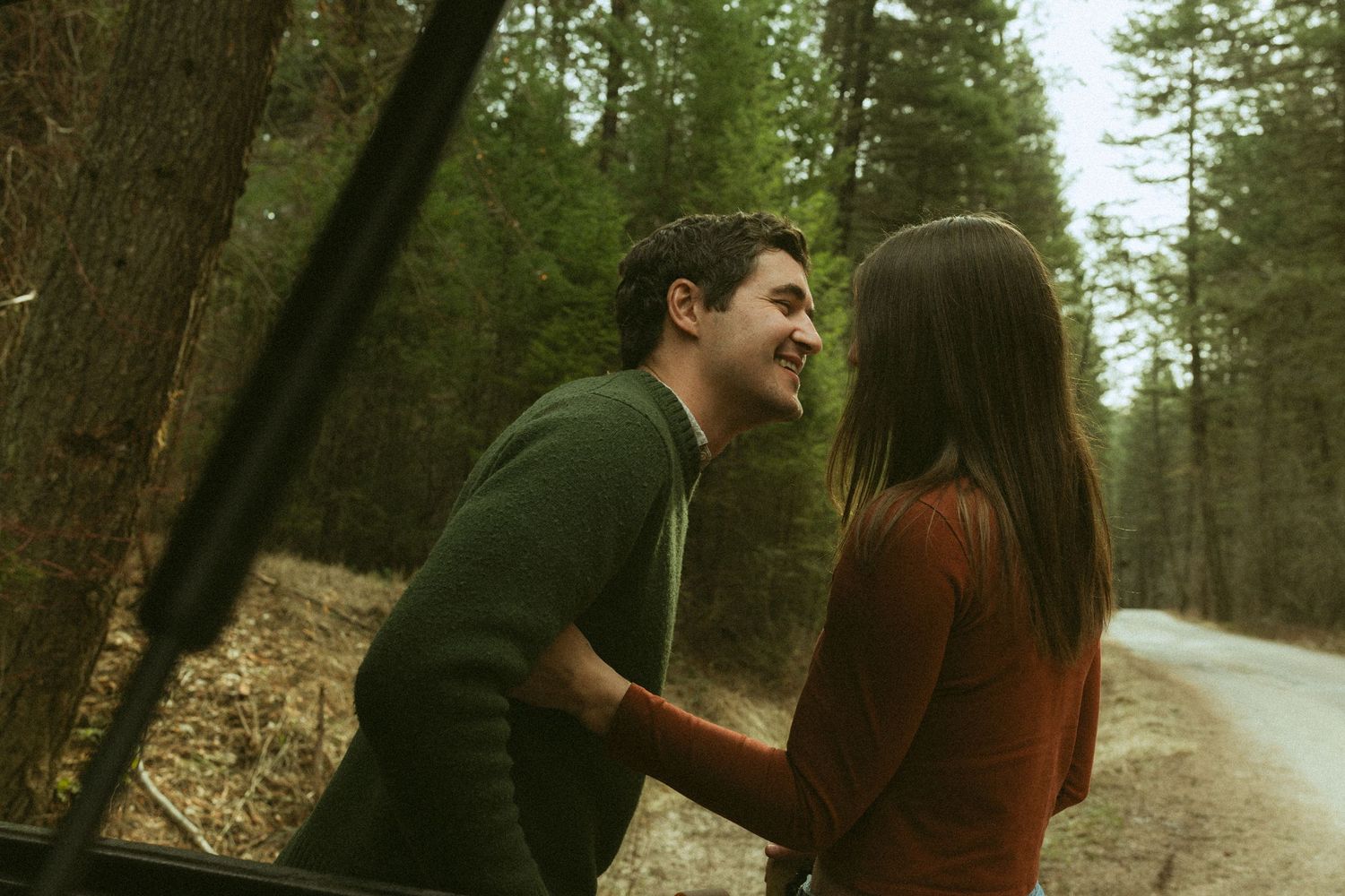 A smiling couple shares a romantic moment outdoors near a forest road, with tall trees in the background.