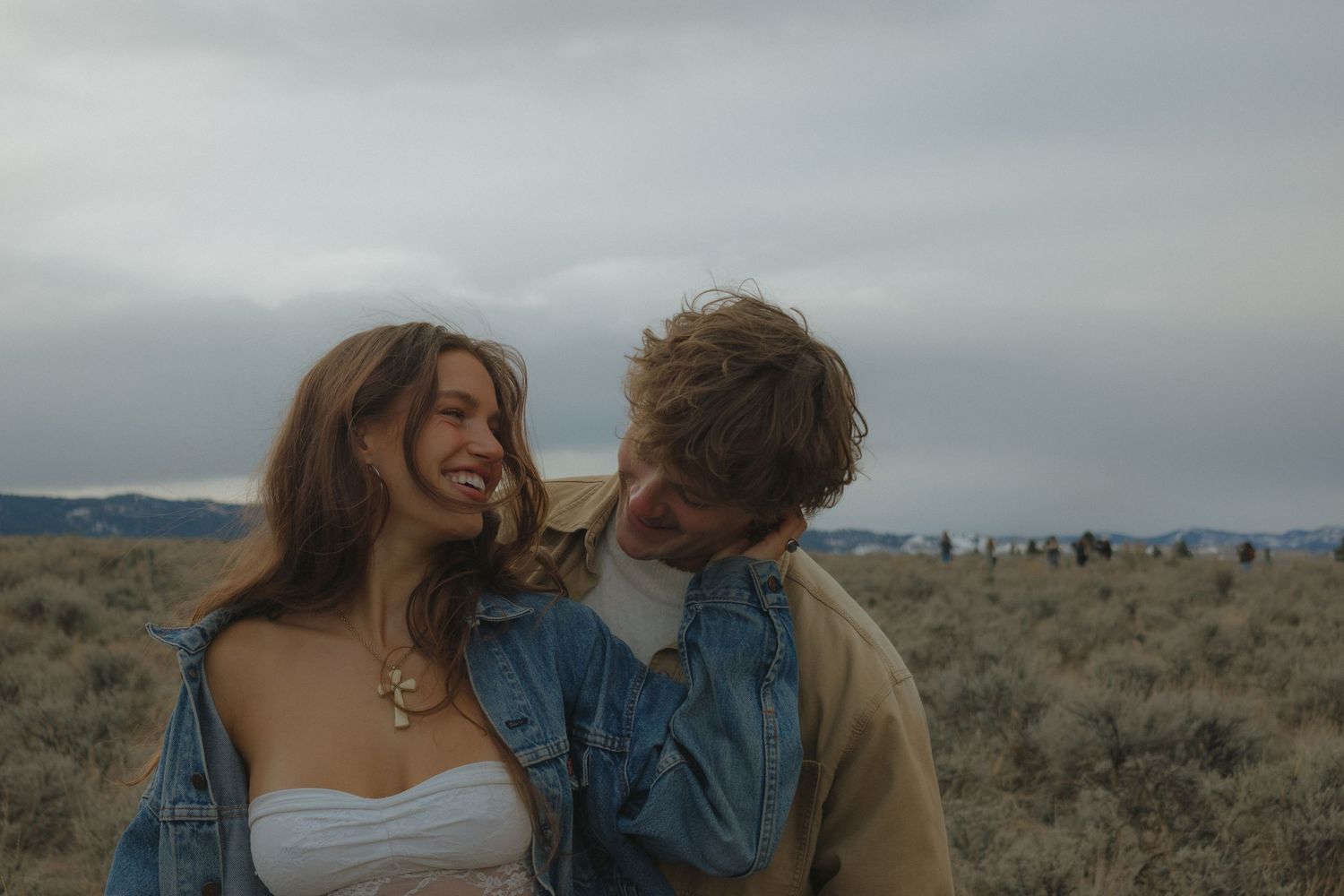 Couple embracing in a desert landscape with mountains in the background during sunset.