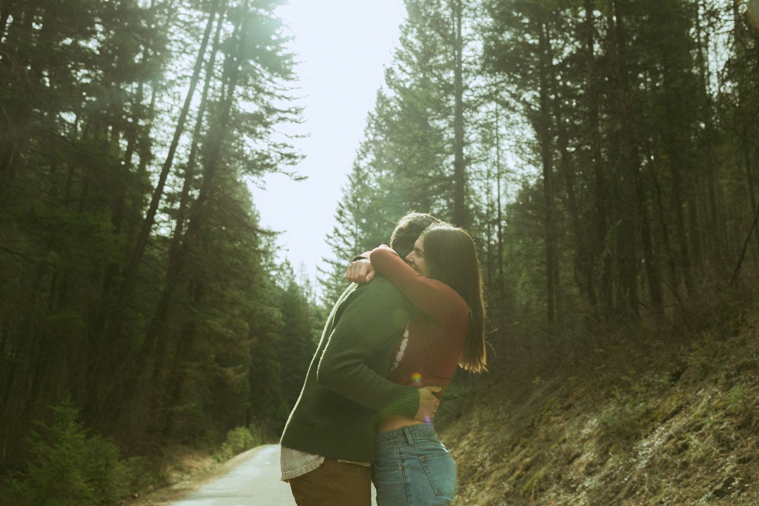 Couple embracing on a scenic forest road surrounded by tall green trees in soft natural light.