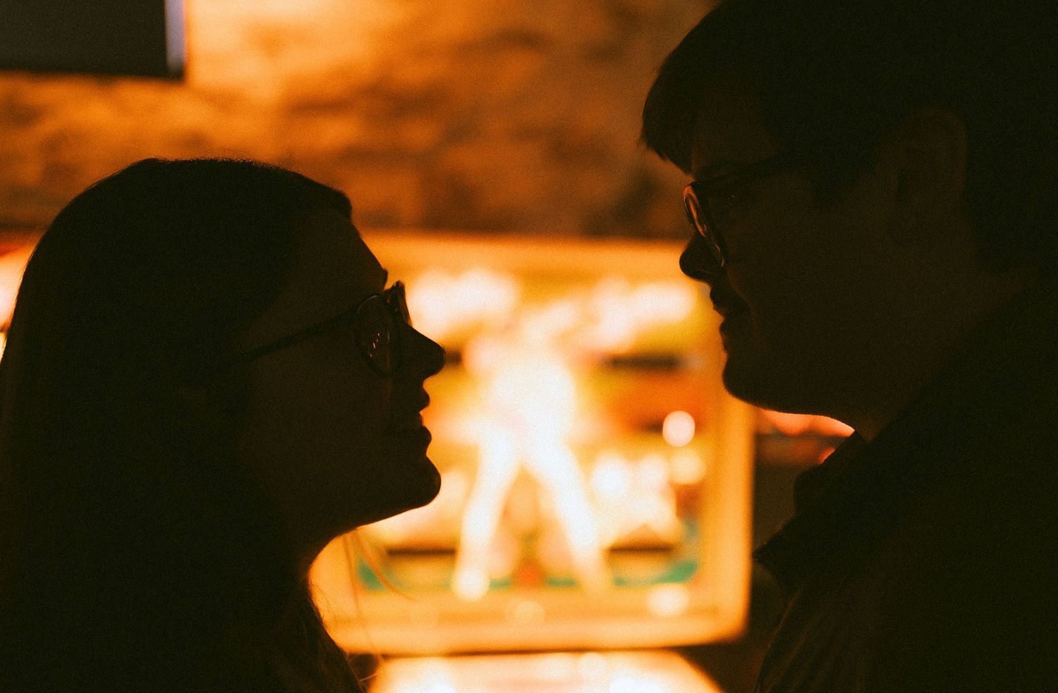 Two silhouettes in profile against warm glowing firelight creating an intimate atmosphere.