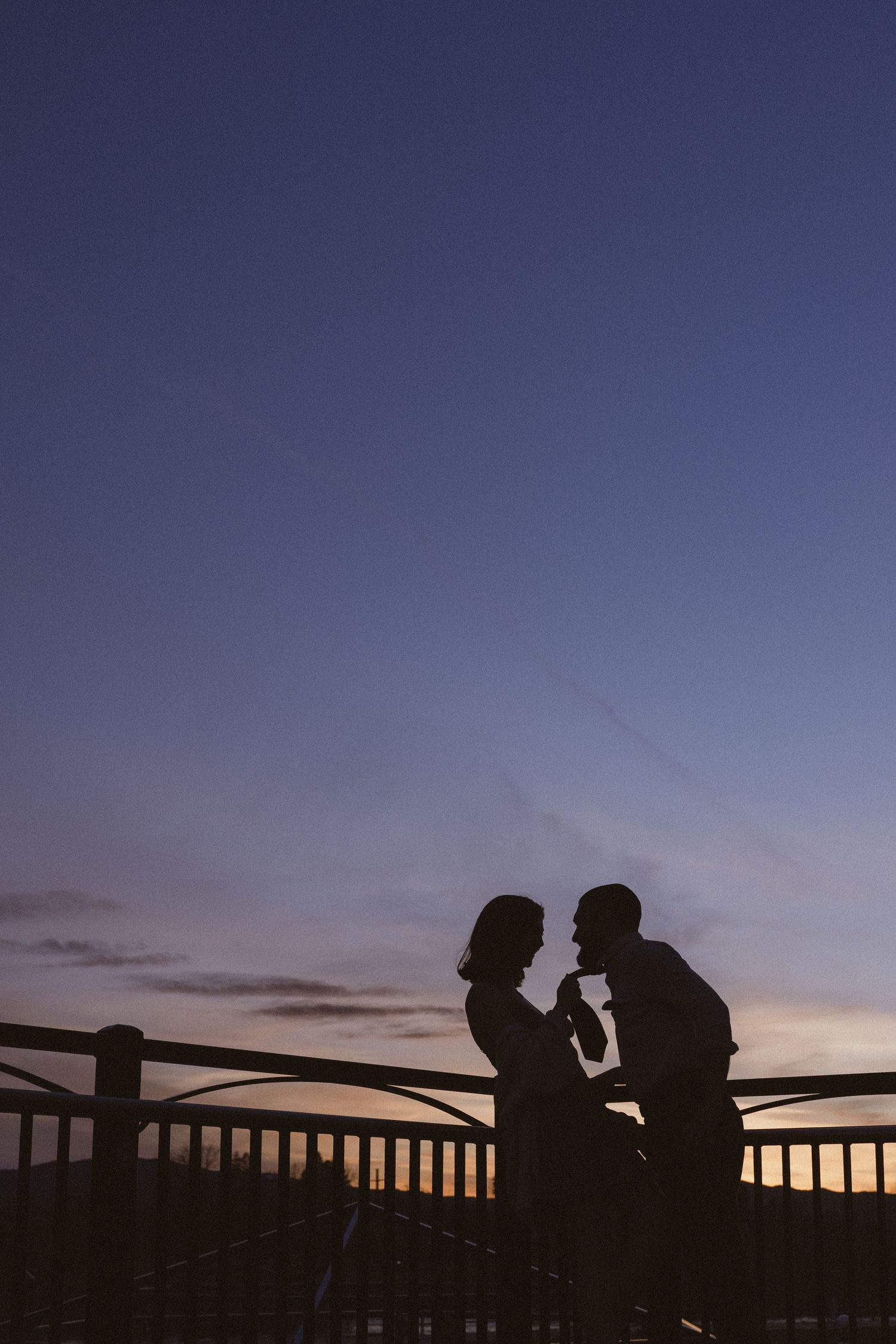Romantic silhouette of a couple facing each other at sunset on a bridge railing with a colorful twilight sky.