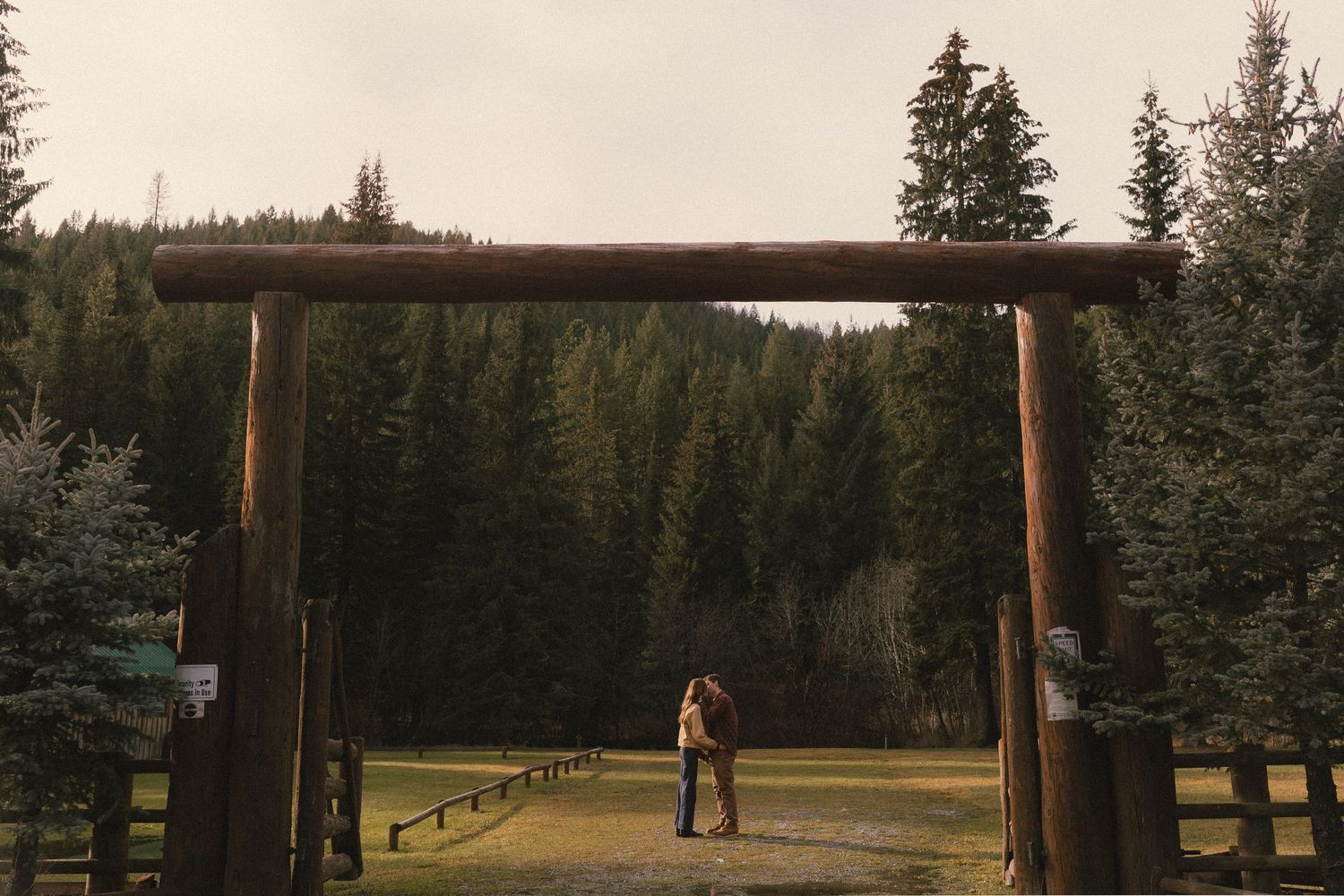A silhouetted figure stands beneath a rustic wooden archway entrance surrounded by evergreen trees at sunset.