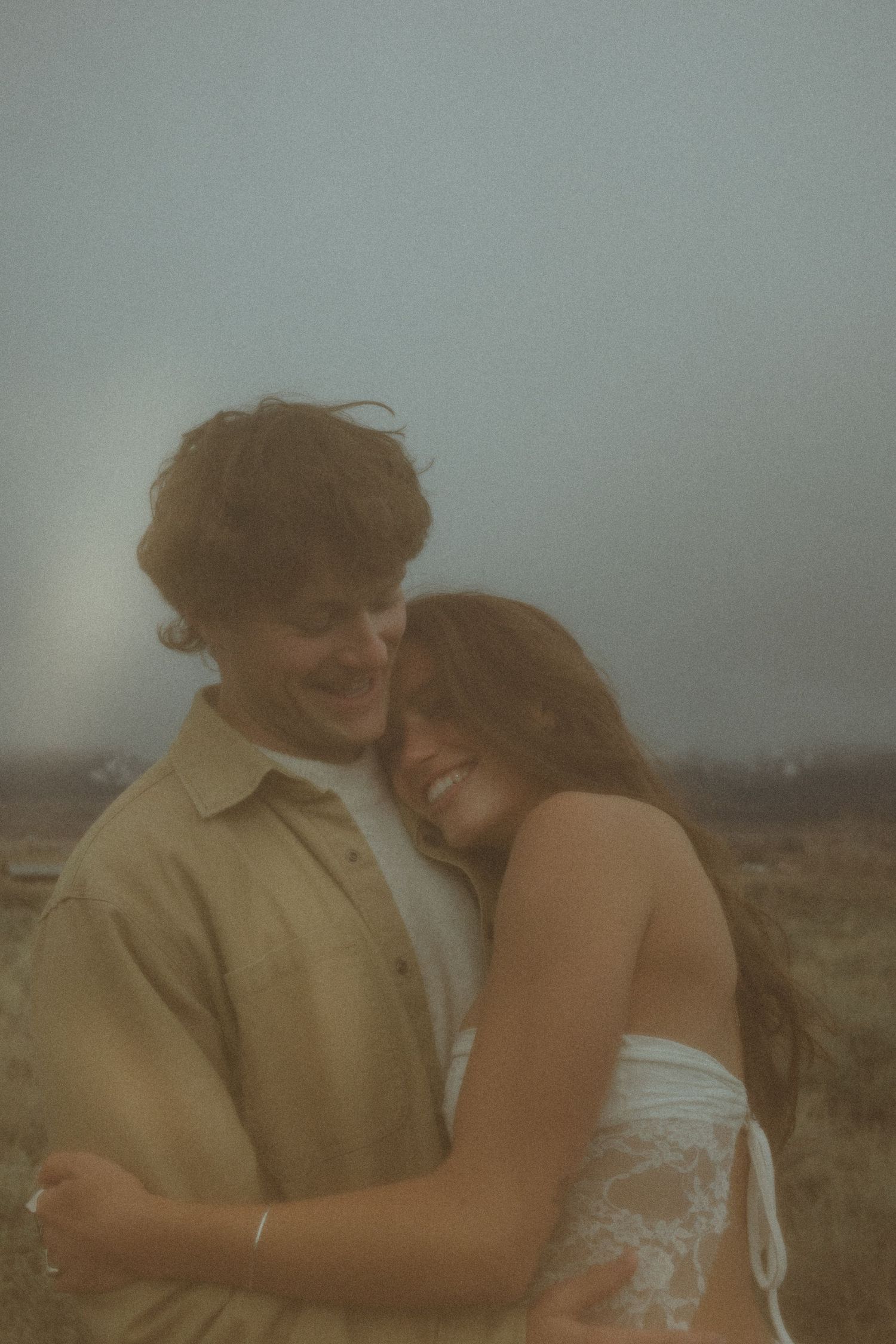 A couple embraces in a dreamy, foggy outdoor setting with muted tones and soft lighting.