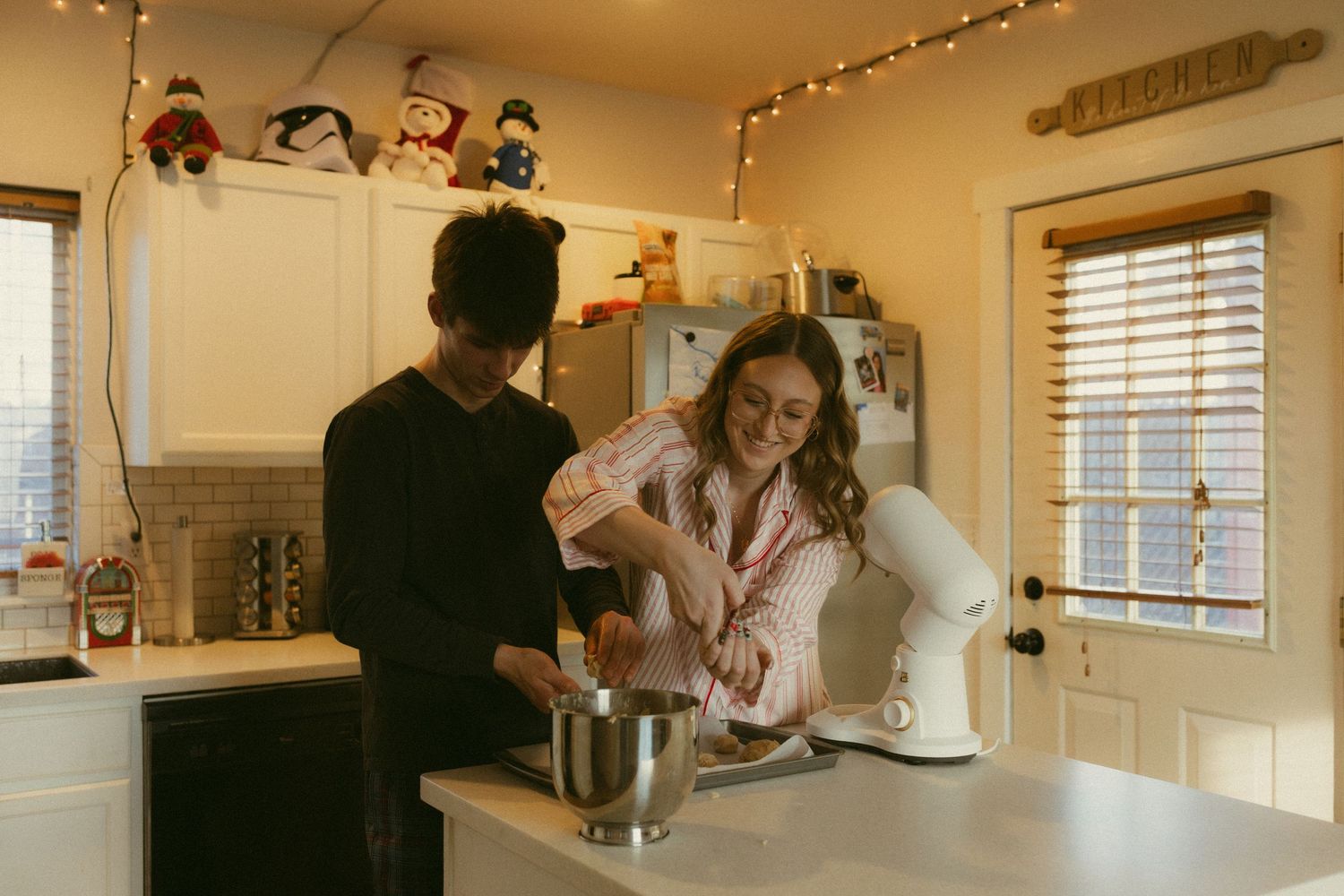 Warm string lights illuminate a cozy kitchen interior with wooden cabinets and windows creating an inviting atmosphere.