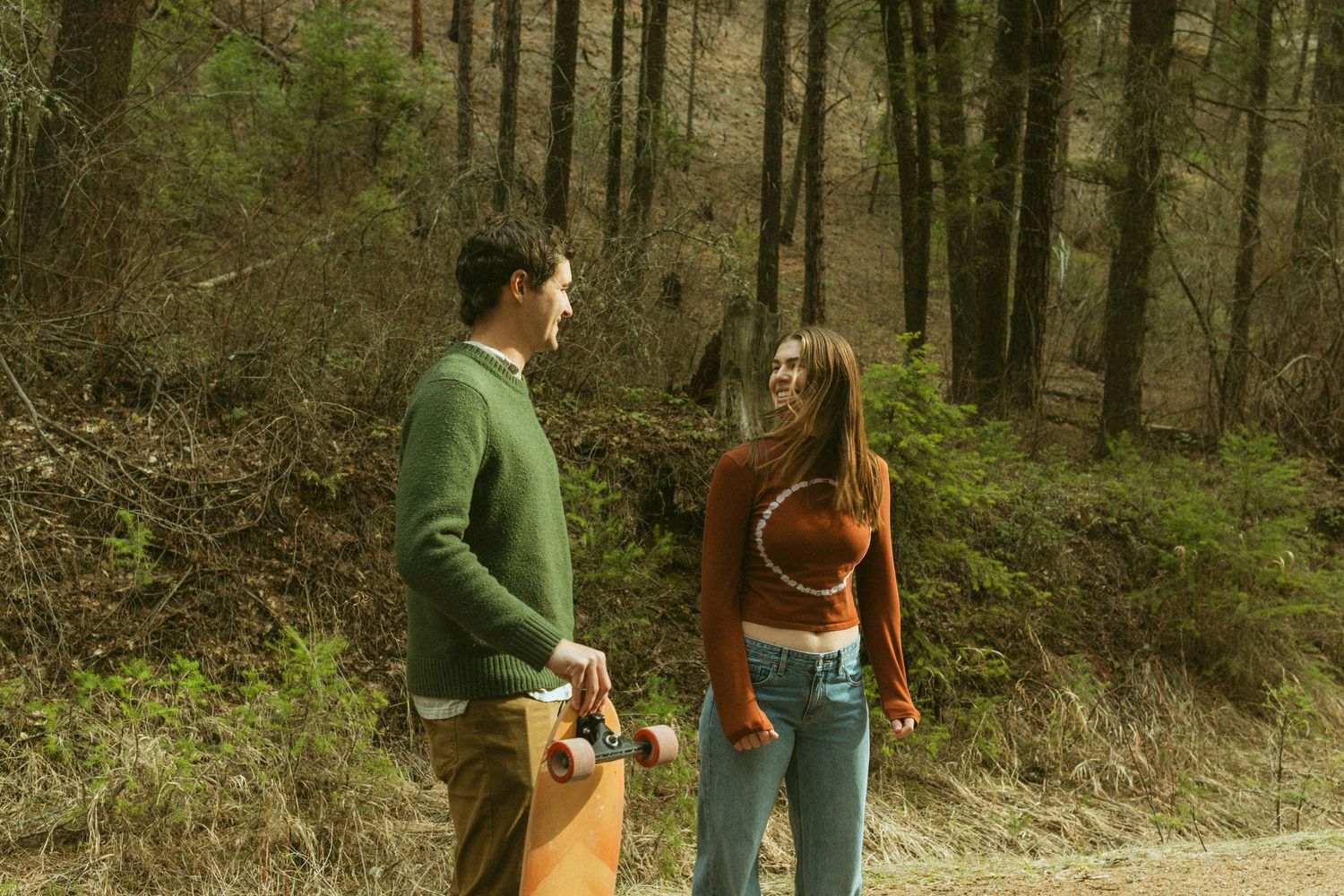 A young couple talks on a forest trail, man holding a skateboard in a green sweater, woman in a rust top and jeans.