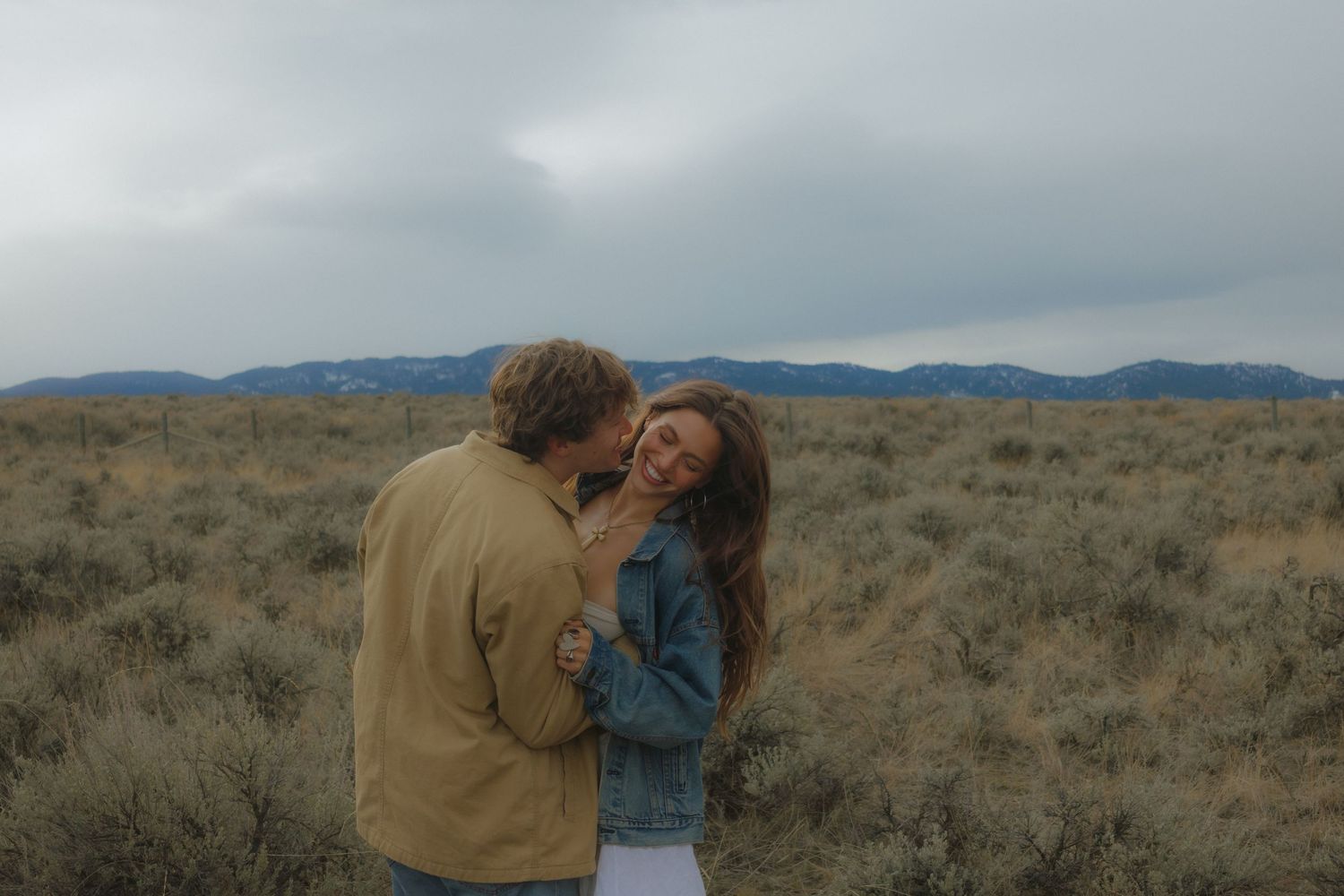 Two people share an intimate moment in a desert landscape with mountains and cloudy skies in the background.