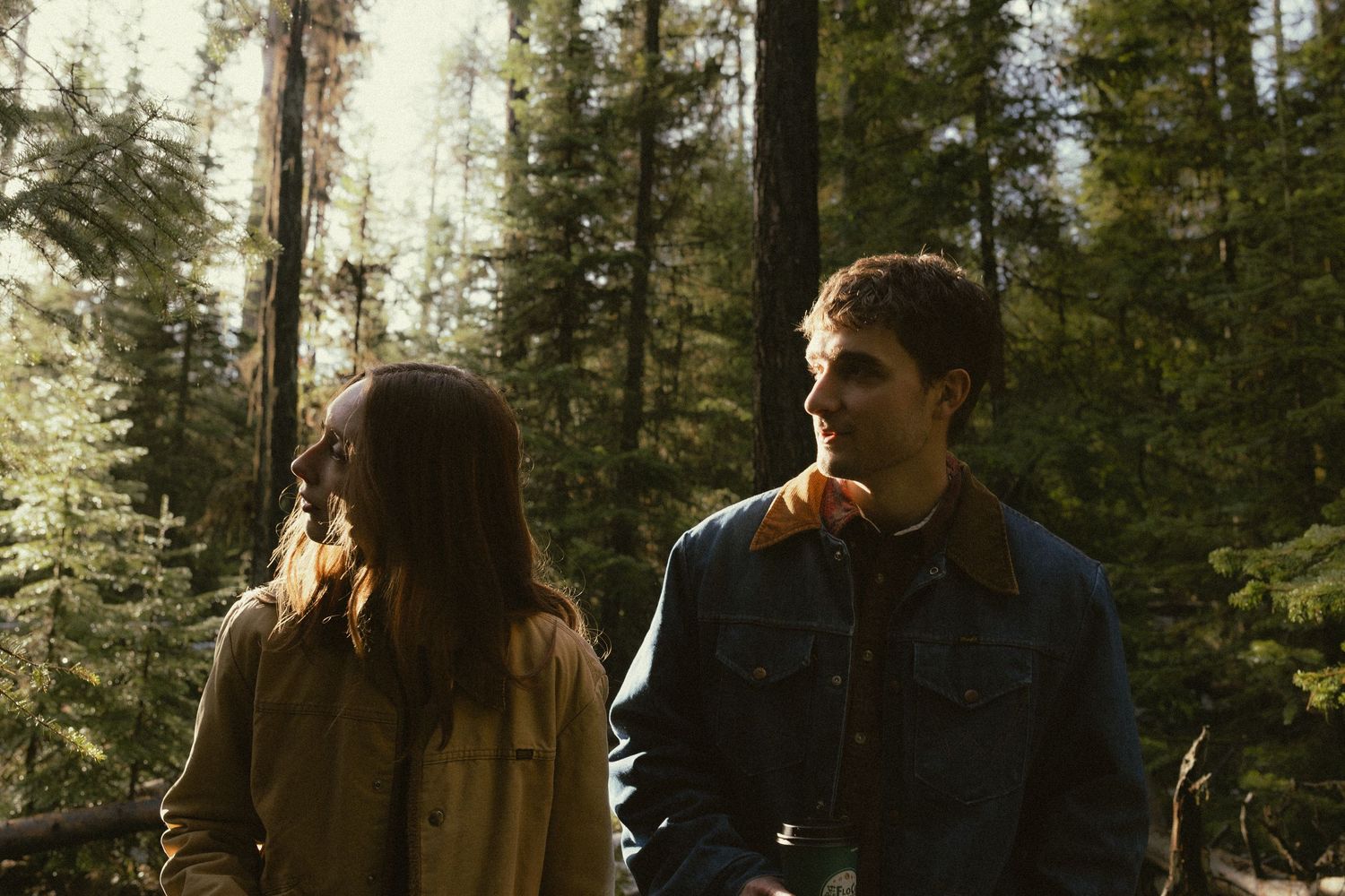 A couple embraces in a romantic moment while standing among tall evergreen trees in a forest setting.