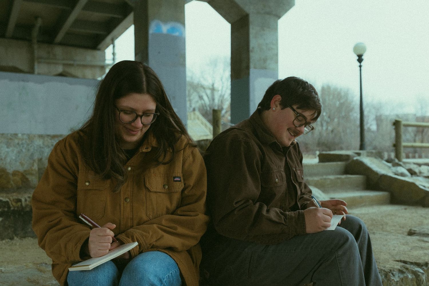Two people sit together on concrete steps while writing in notebooks outdoors.