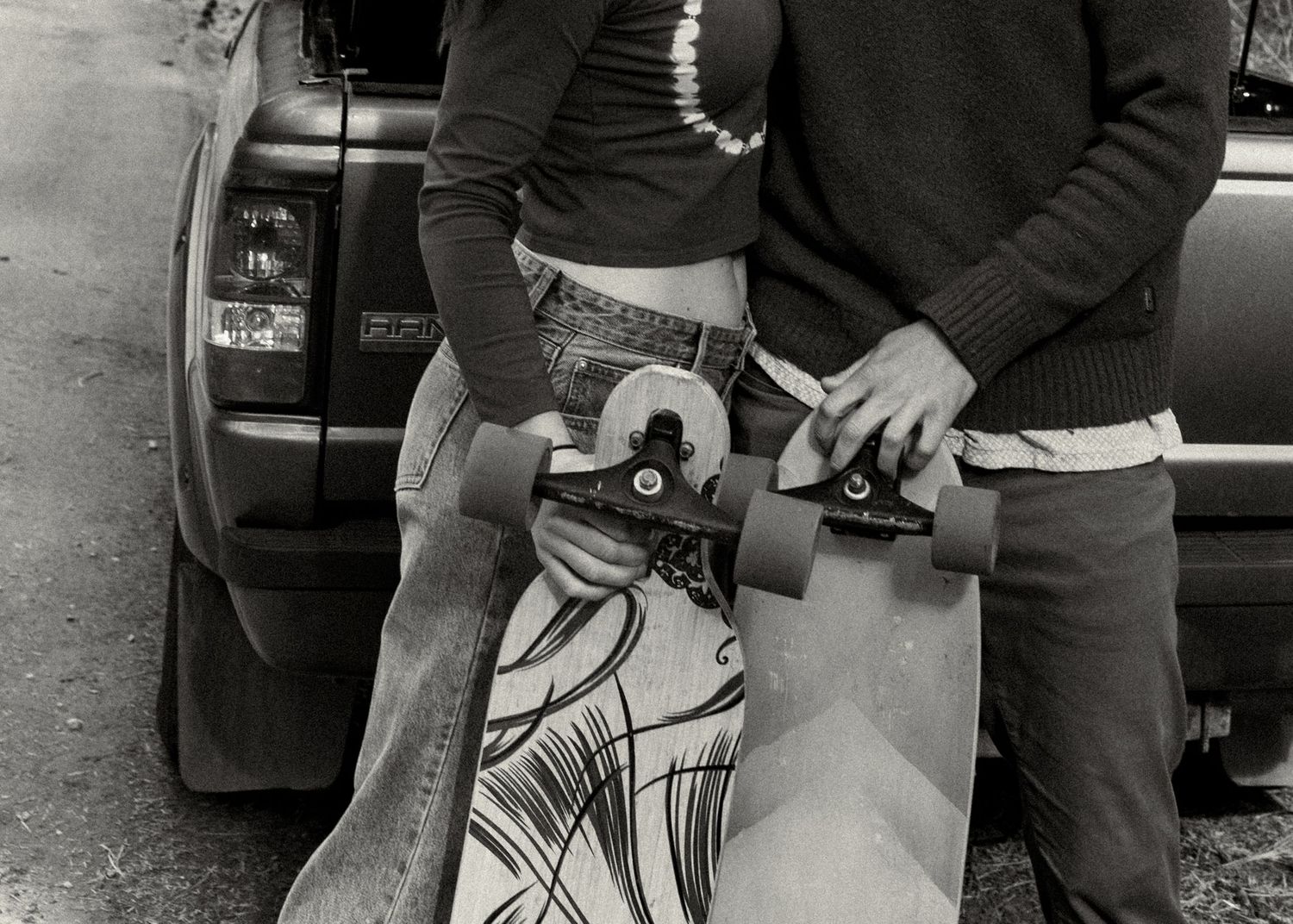 Two people holding a longboard skateboard together in black and white photo near a parked SUV.