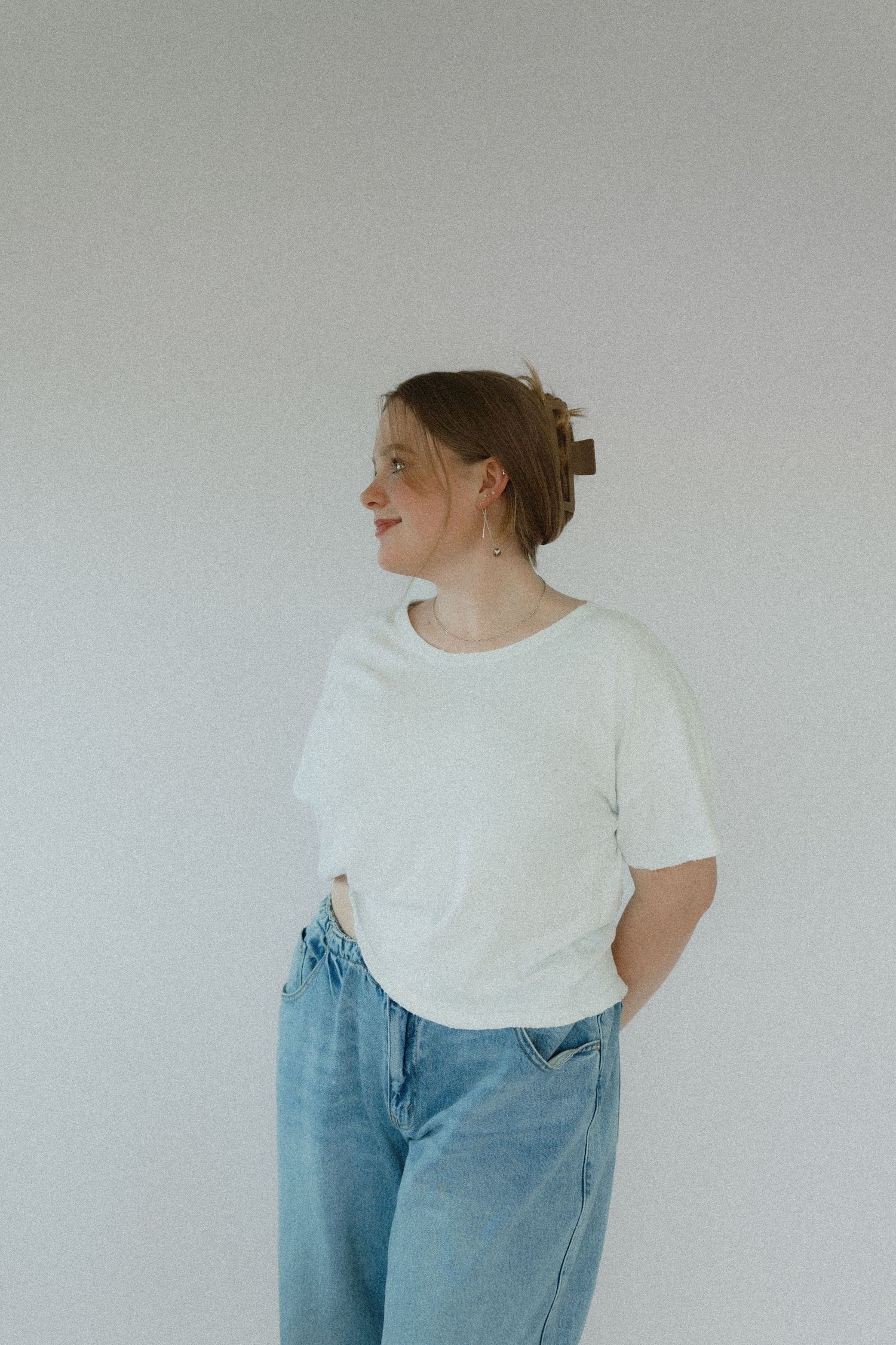 A figure in a casual white t-shirt and light blue jeans stands against a plain white wall looking to the side.