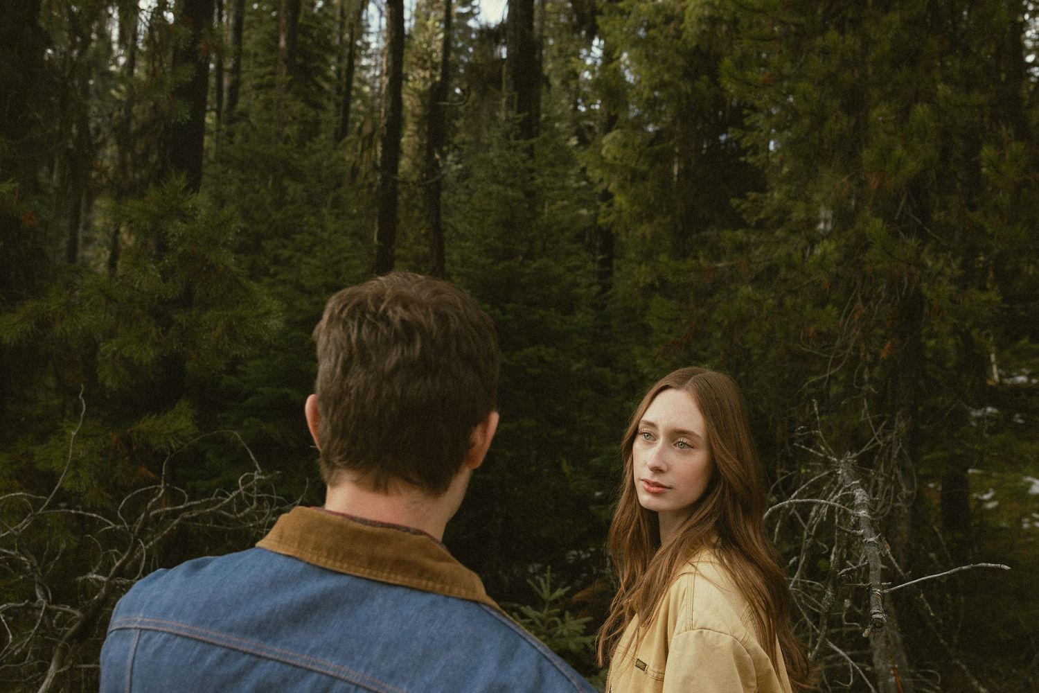 A couple walks together through a lush green forest during golden hour with sunlight filtering through the trees.