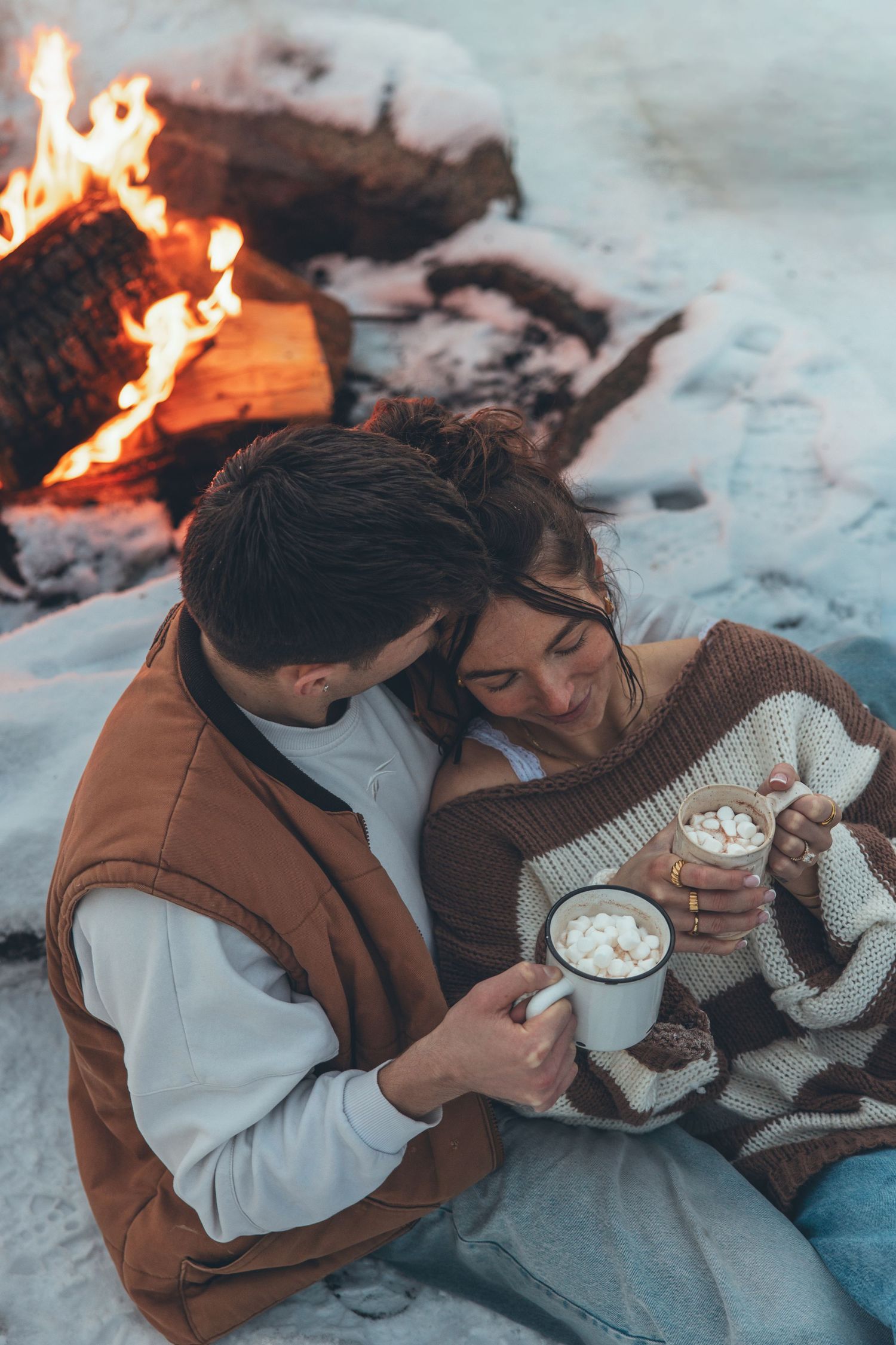 A couple cuddling by a campfire with hot drinks and blankets in the snow.