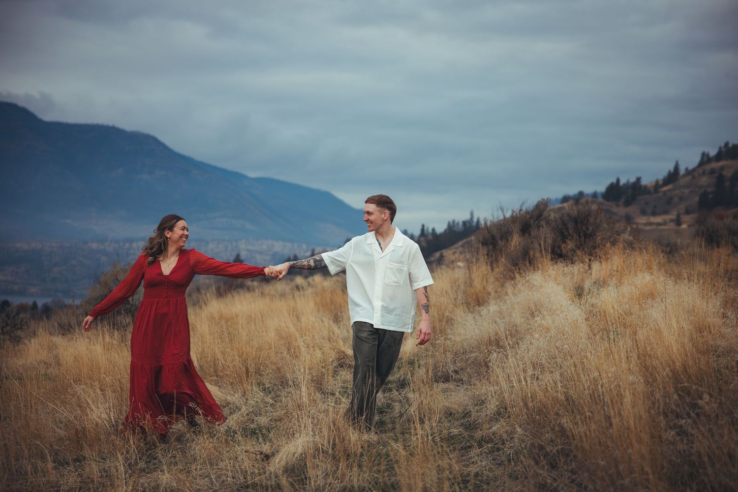 Couple in red dress and white shirt pose dramatically in mountain meadow.