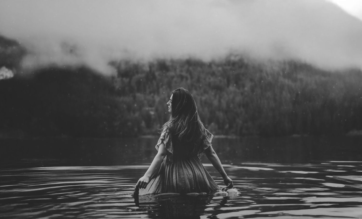 Silhouette of person standing in lake with mountains and fog in background.