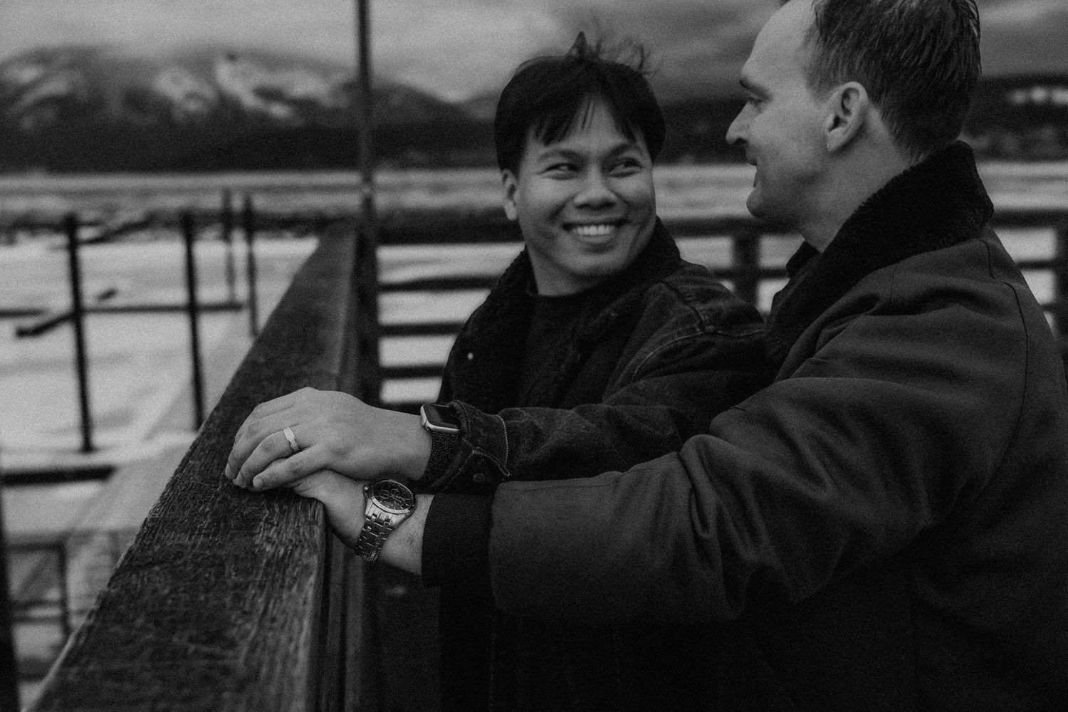 Two people sharing a joyful moment while leaning on a railing with snowy mountains in the background.