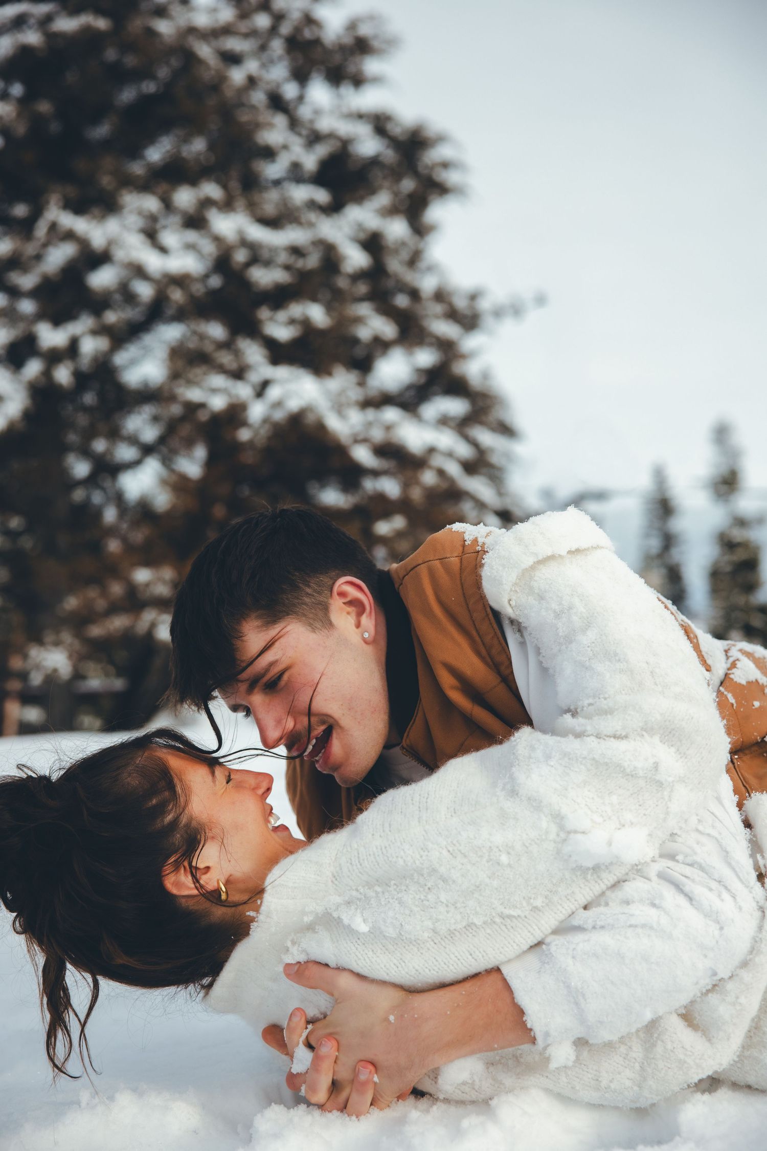 A couple embracing in the snow wearing white and brown winter outfits.