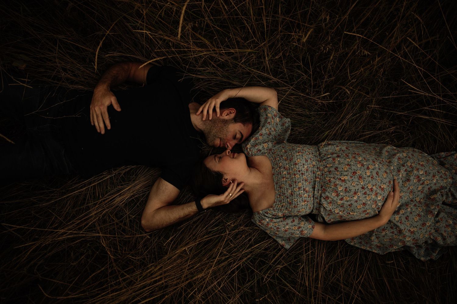 Romantic couple lying in tall grass at sunset captured in moody dark tones.