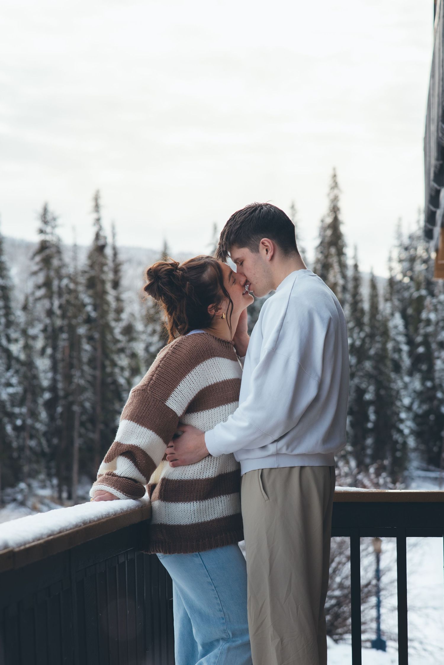 A couple sharing an intimate moment on a snowy balcony with evergreen trees in the background.