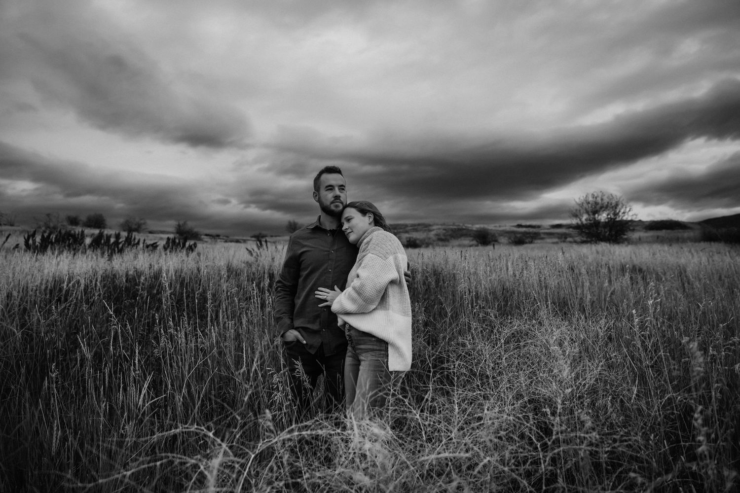 A couple stands together in a golden wheat field under dramatic cloudy skies at sunset.