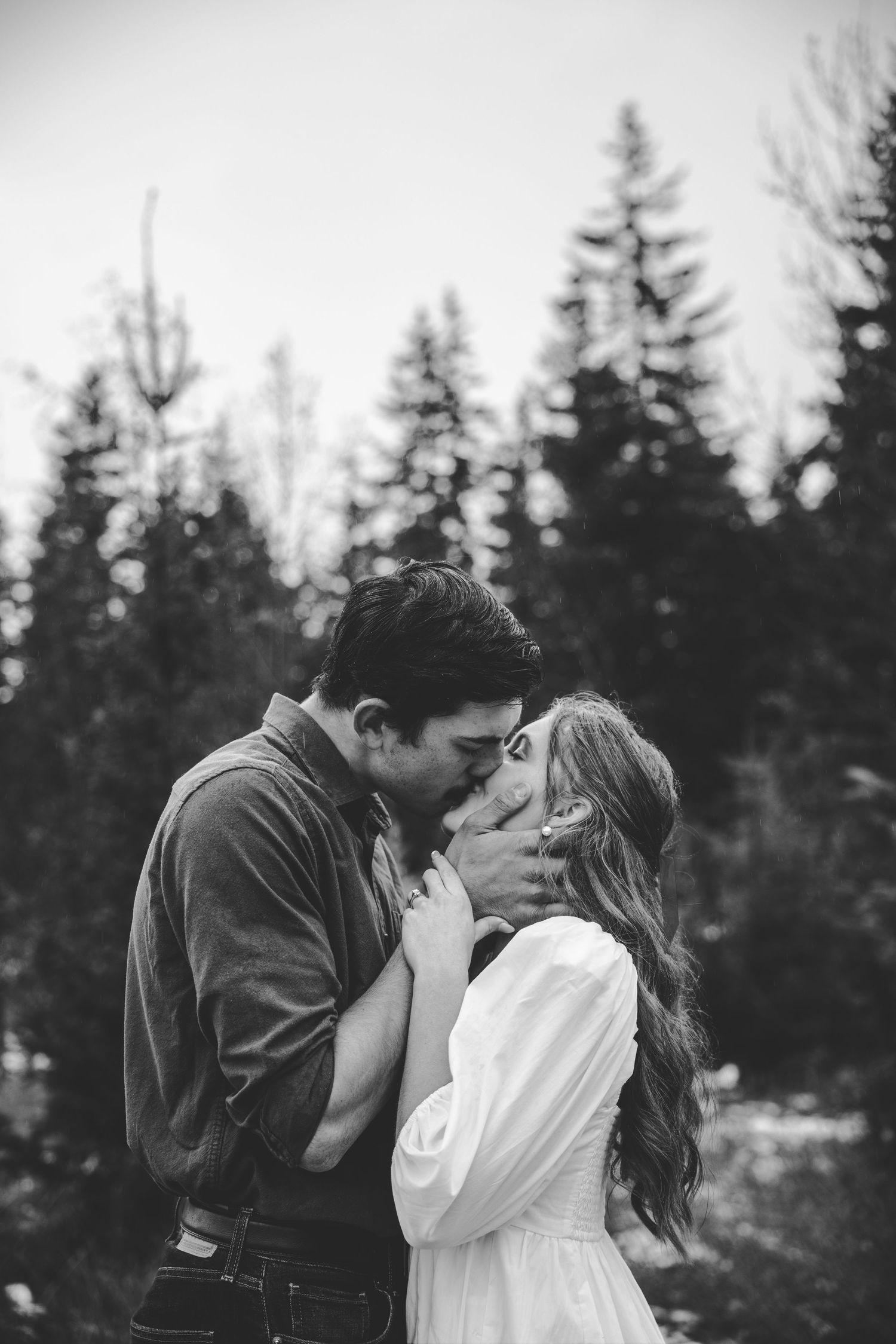 Black and white photograph of a romantic moment with evergreen trees silhouetted in the background.
