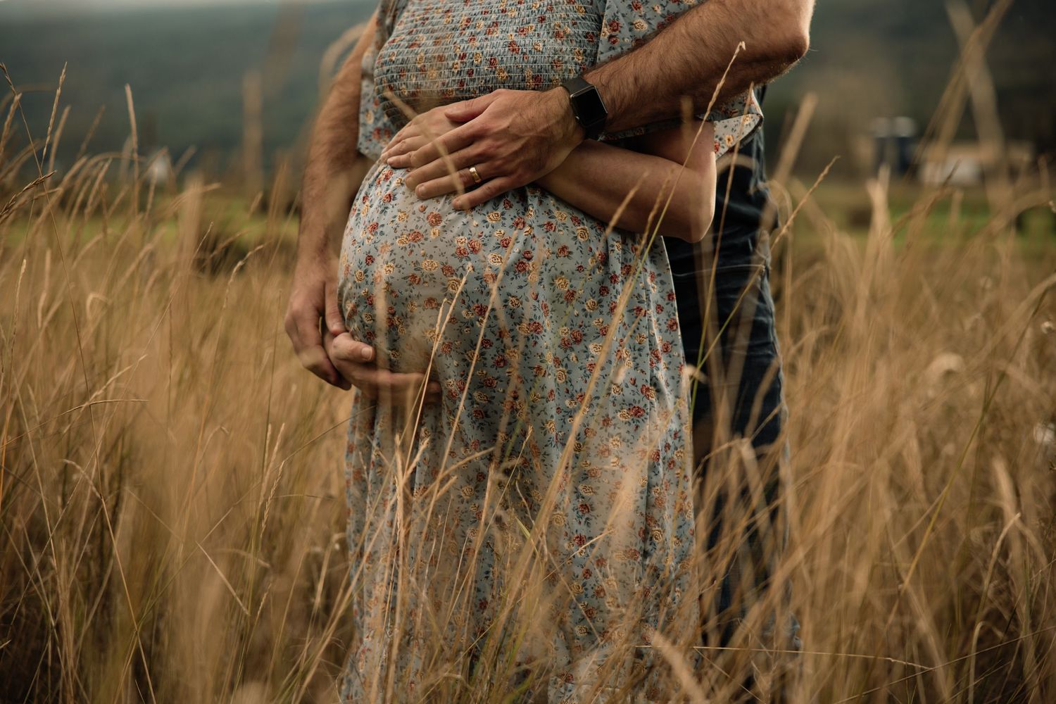 A couple stands close together in a field of tall golden grass during golden hour.