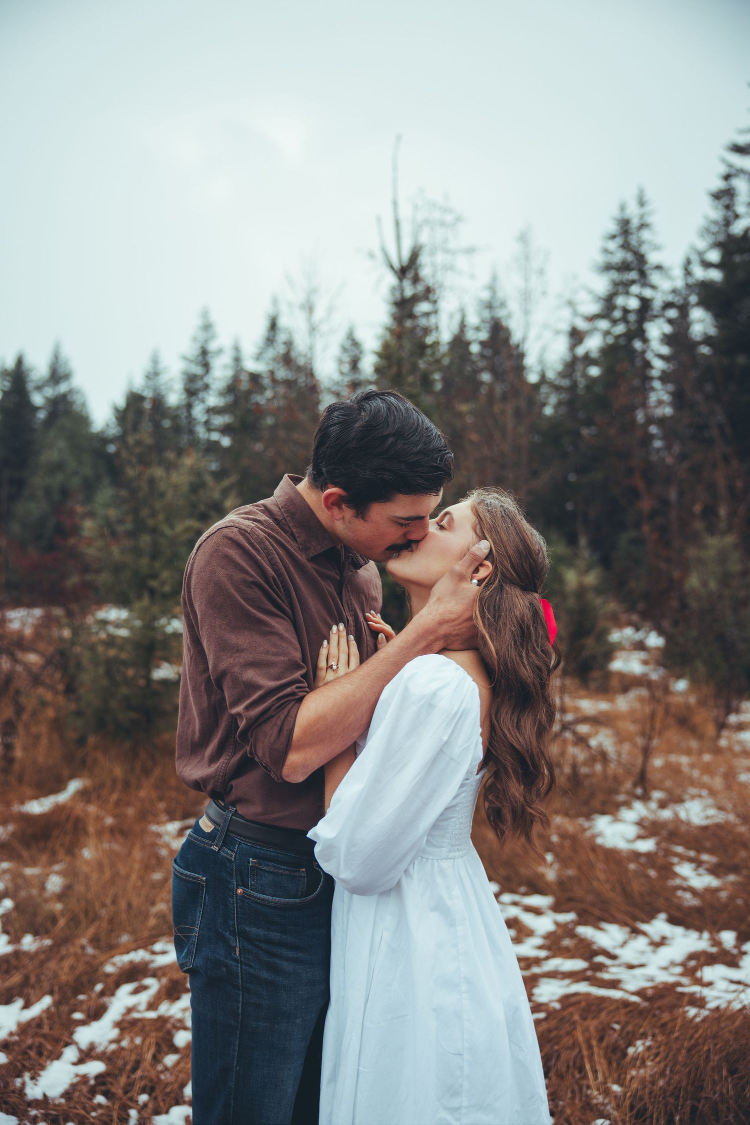 A couple shares a romantic moment in a snowy forest landscape with pine trees.