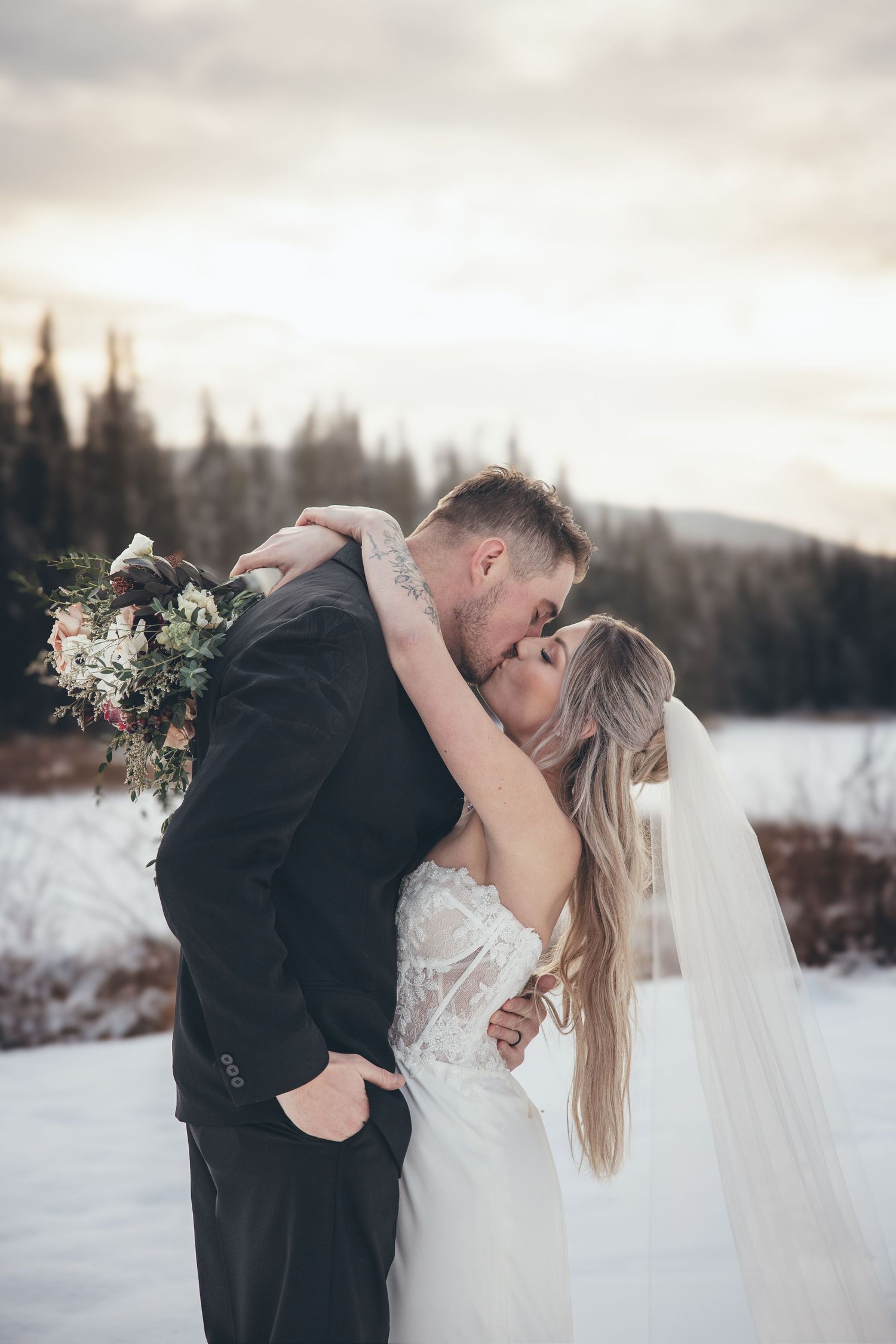 Romantic winter wedding portrait with couple sharing intimate kiss in snowy mountain setting.