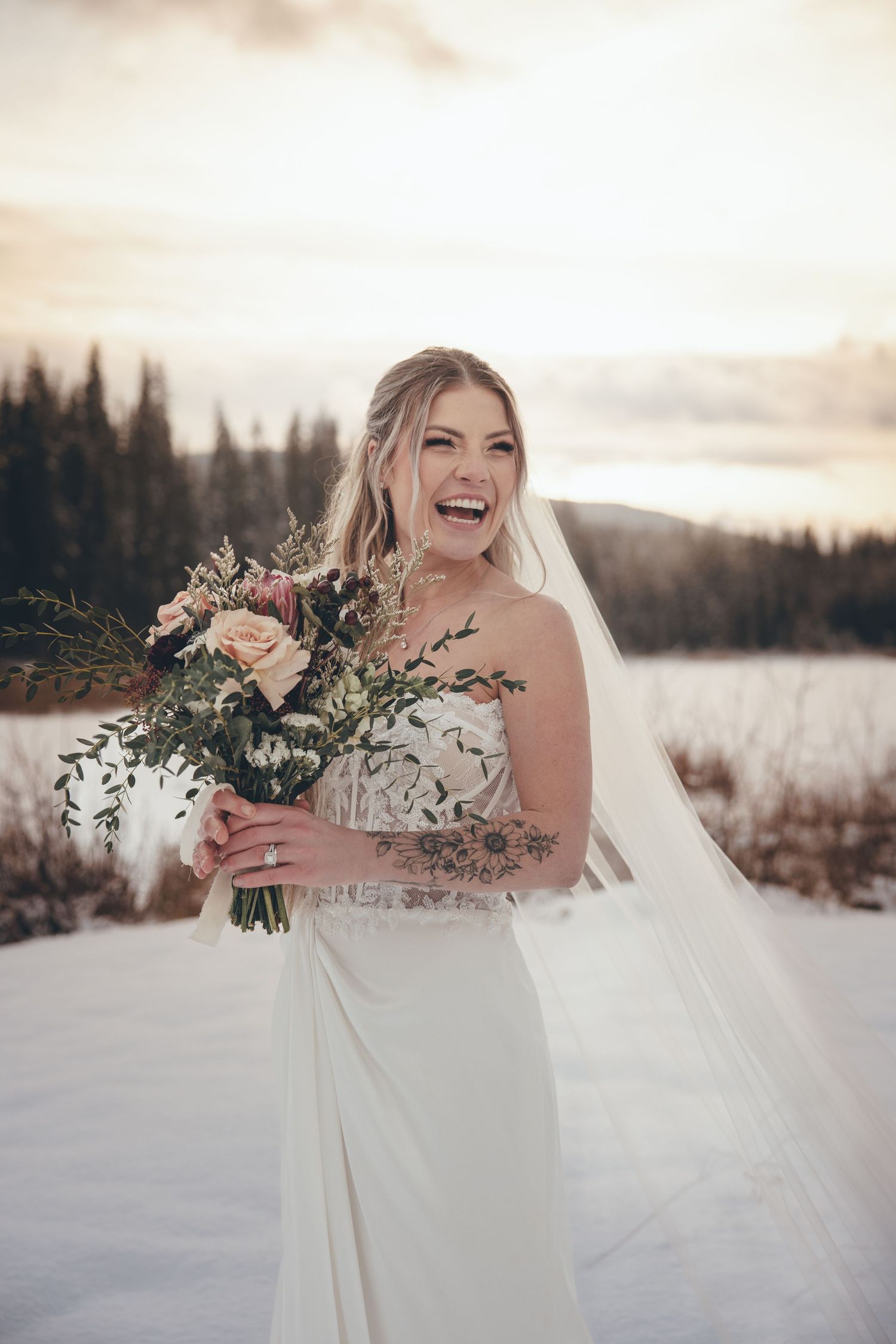 Winter bride in white dress holding bouquet poses joyfully in snowy landscape with pine trees.