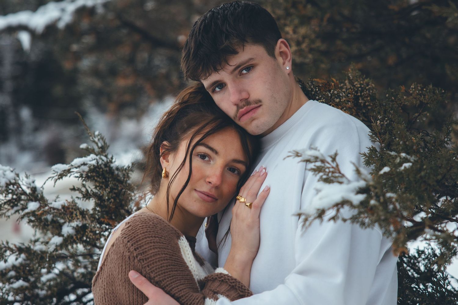 A couple in winter attire embracing among snow-covered trees and branches.