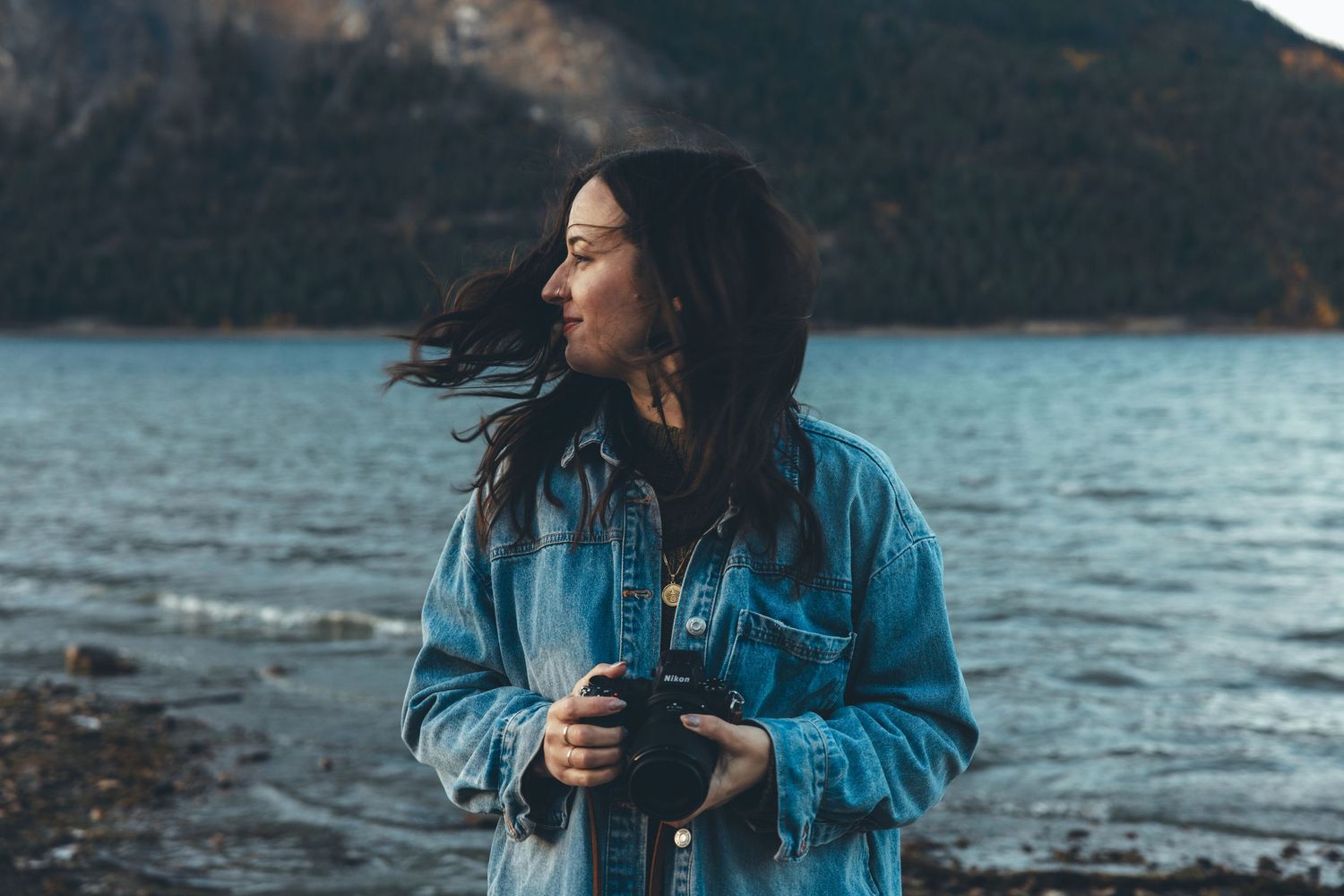 Person in blue denim jacket standing by lake with mountains in background.