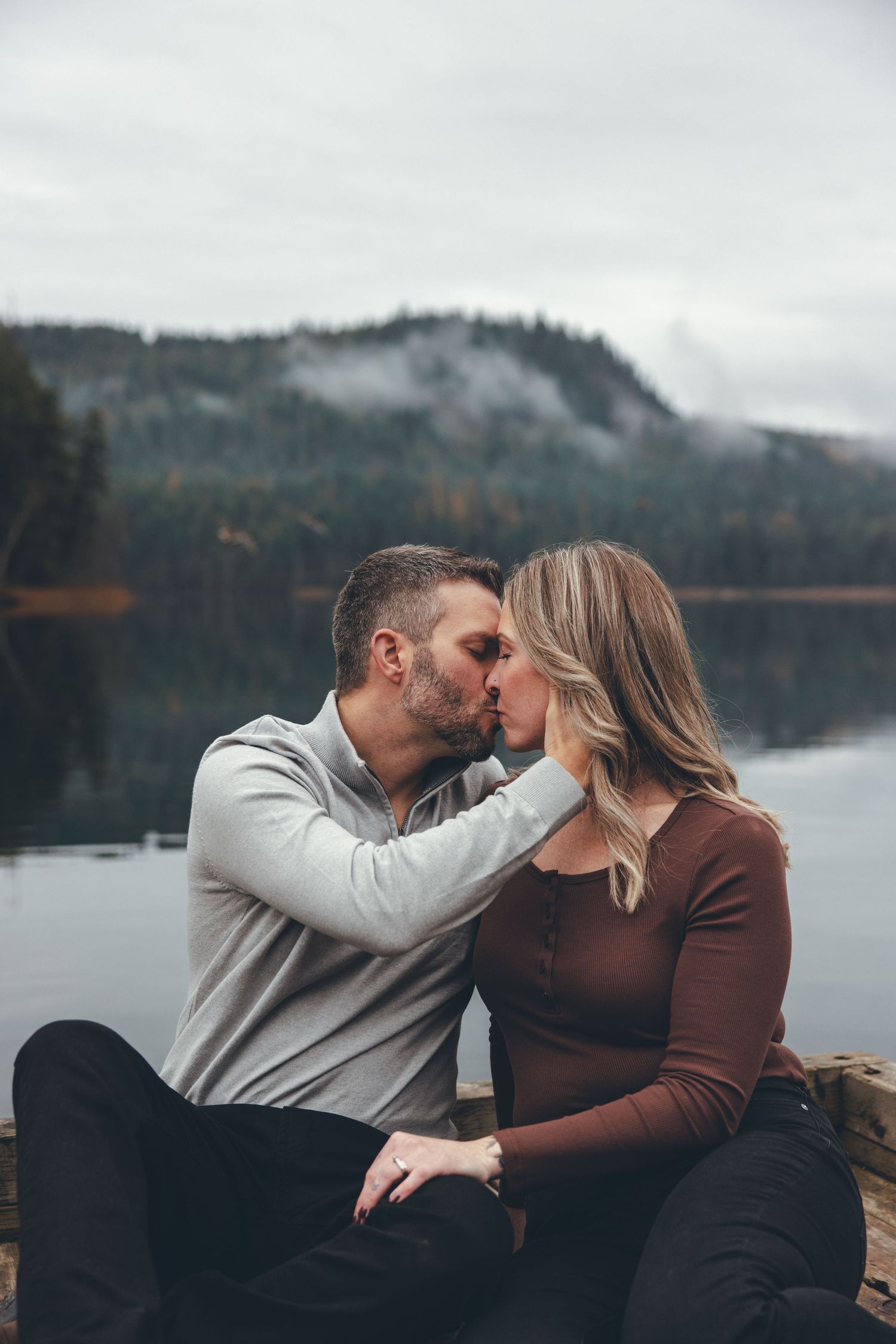 Couple sharing intimate moments on dock with misty mountain lake backdrop.