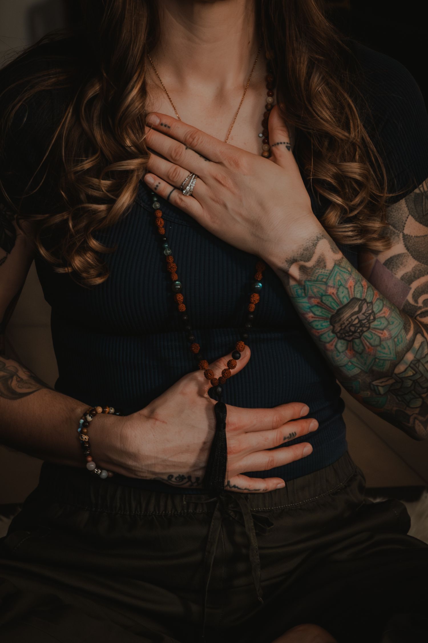 Artistic photo of hands with tattoos and beaded necklace against dark background.