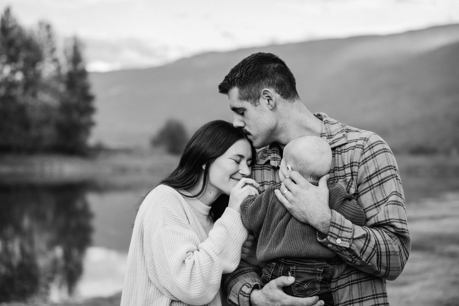 A tender family moment captured outdoors with a baby against a scenic mountain backdrop.