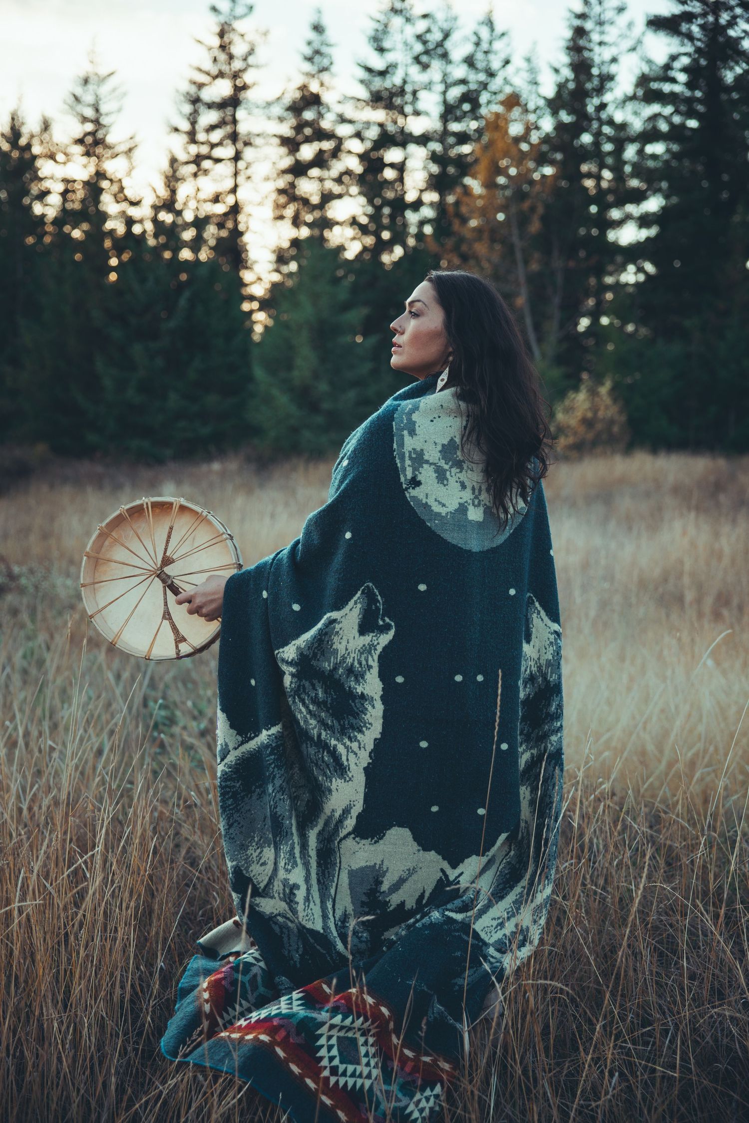 Figure in patterned clothing holding a drum in a field of tall grass at sunset.