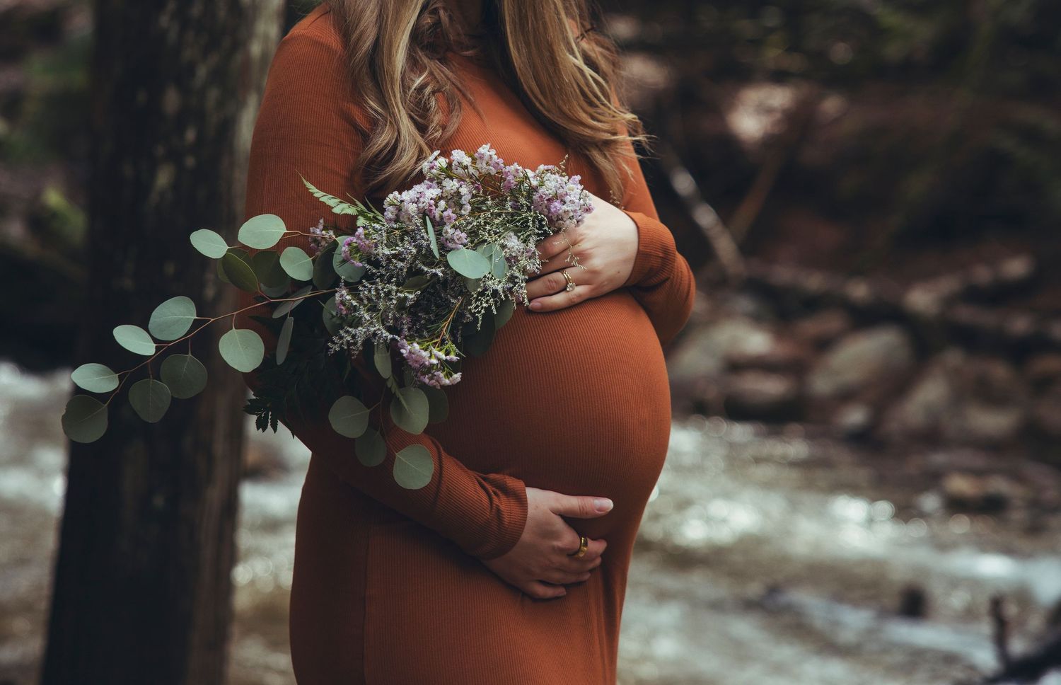 Expectant mother in rust-colored dress holds flowers by a creek in autumn woods.