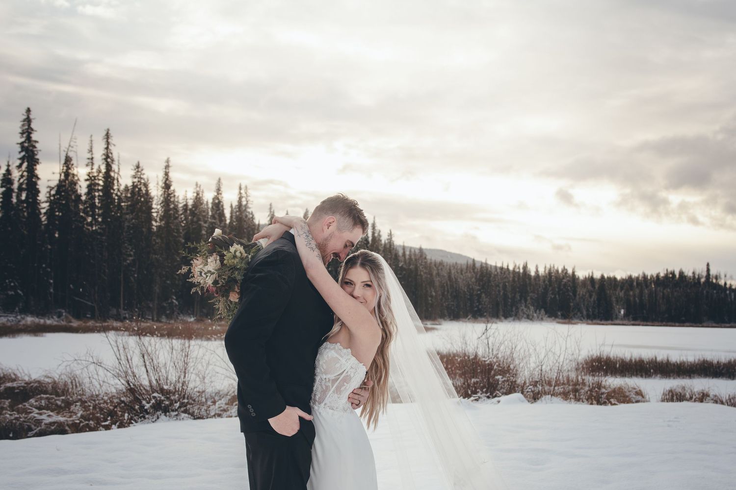 Winter wedding photograph with couple embracing by frozen lake and pine forest.
