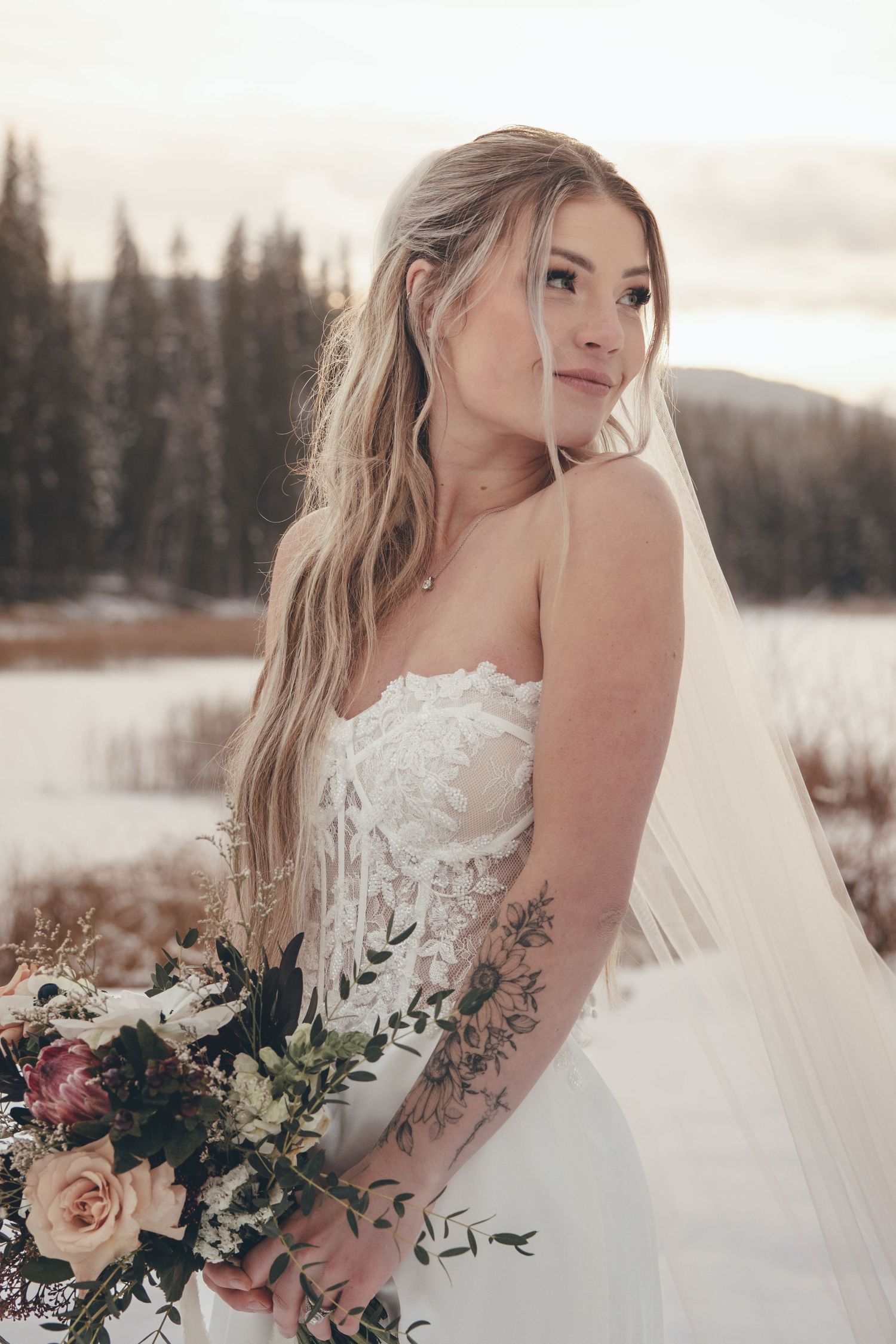 Bride in lace wedding dress poses with bouquet against snowy winter landscape.