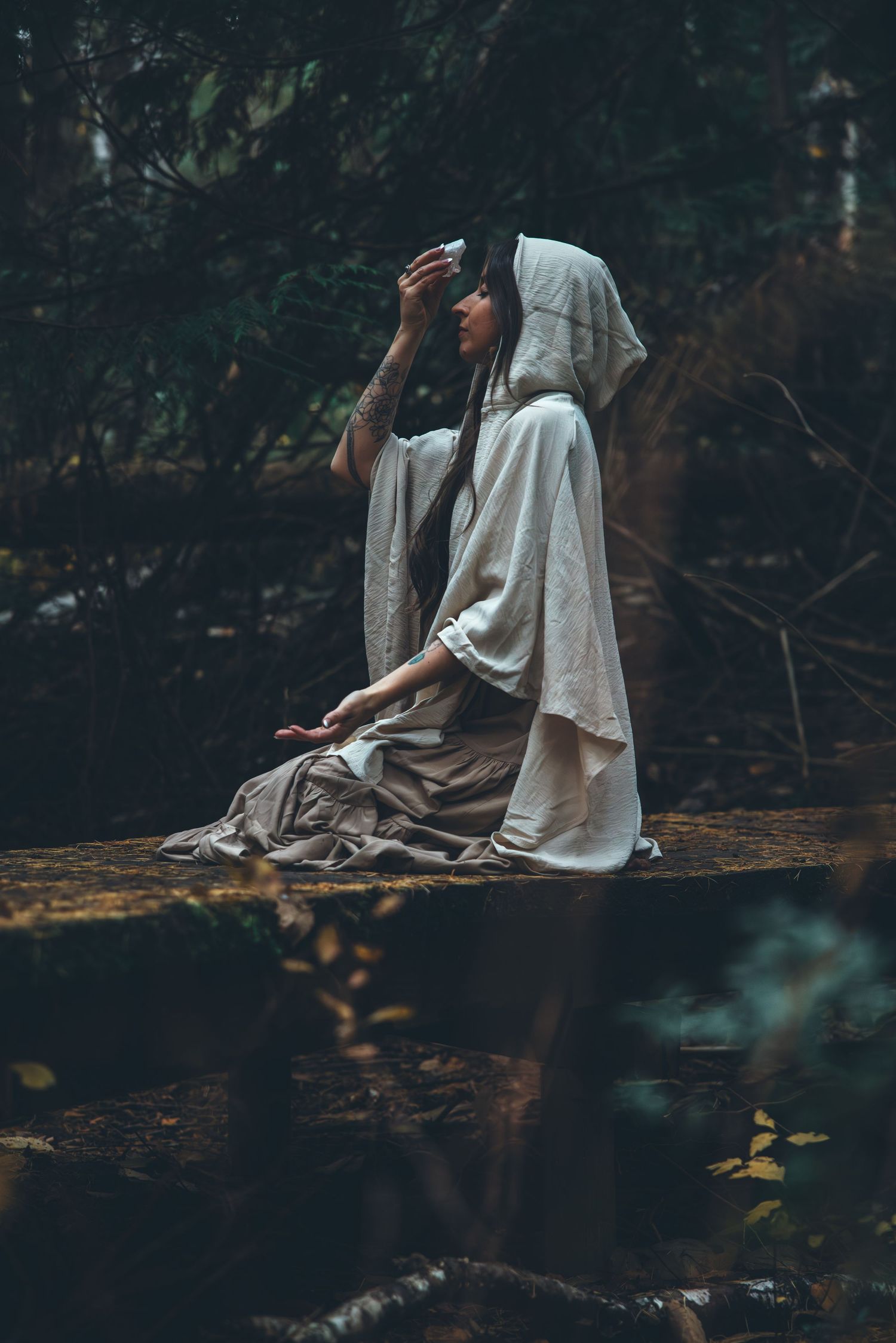 Ethereal scene of someone in a flowing white dress sitting on a log in a dark forest setting.