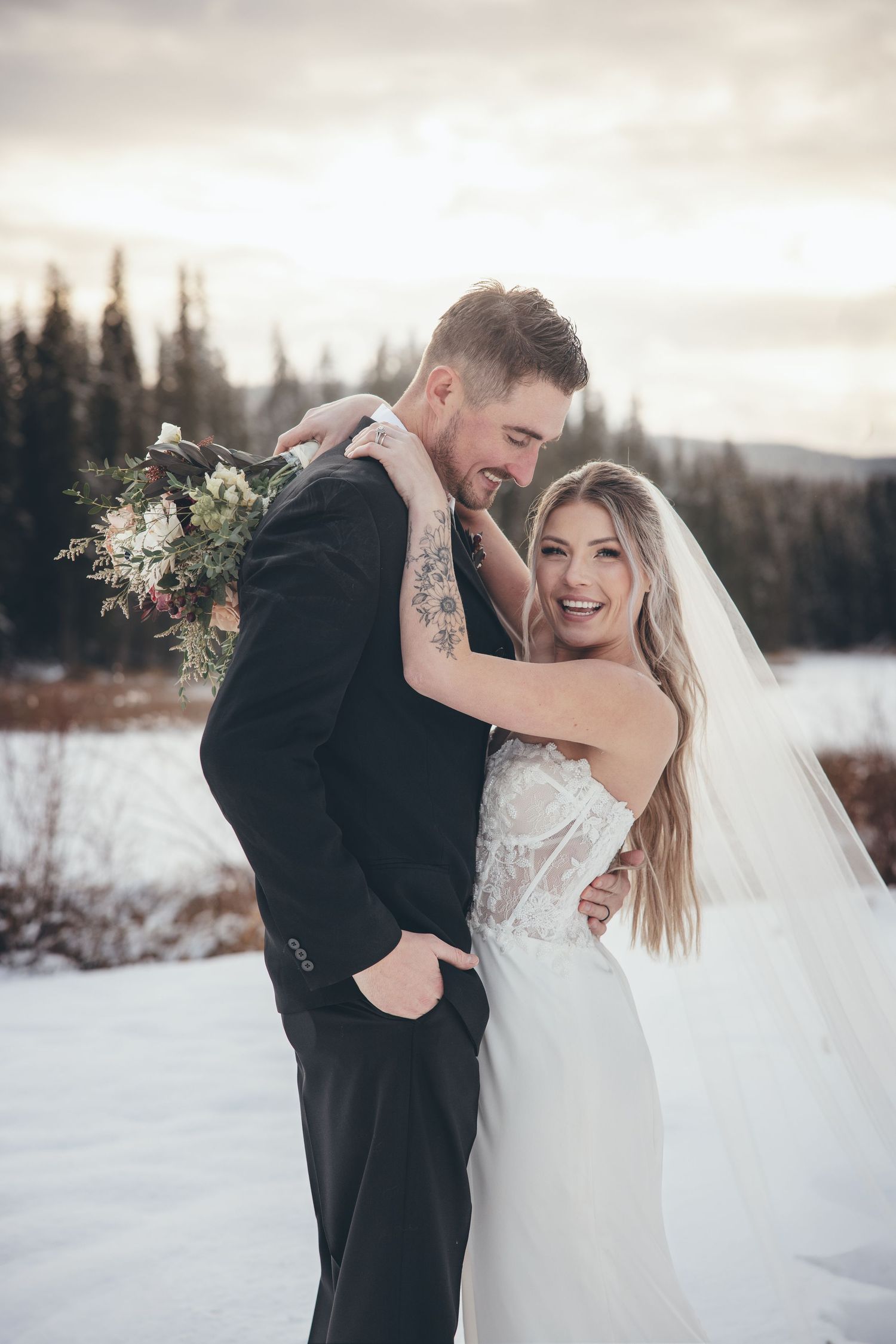 A romantic winter wedding photoshoot with a couple laughing together in the snow with evergreen trees in background.