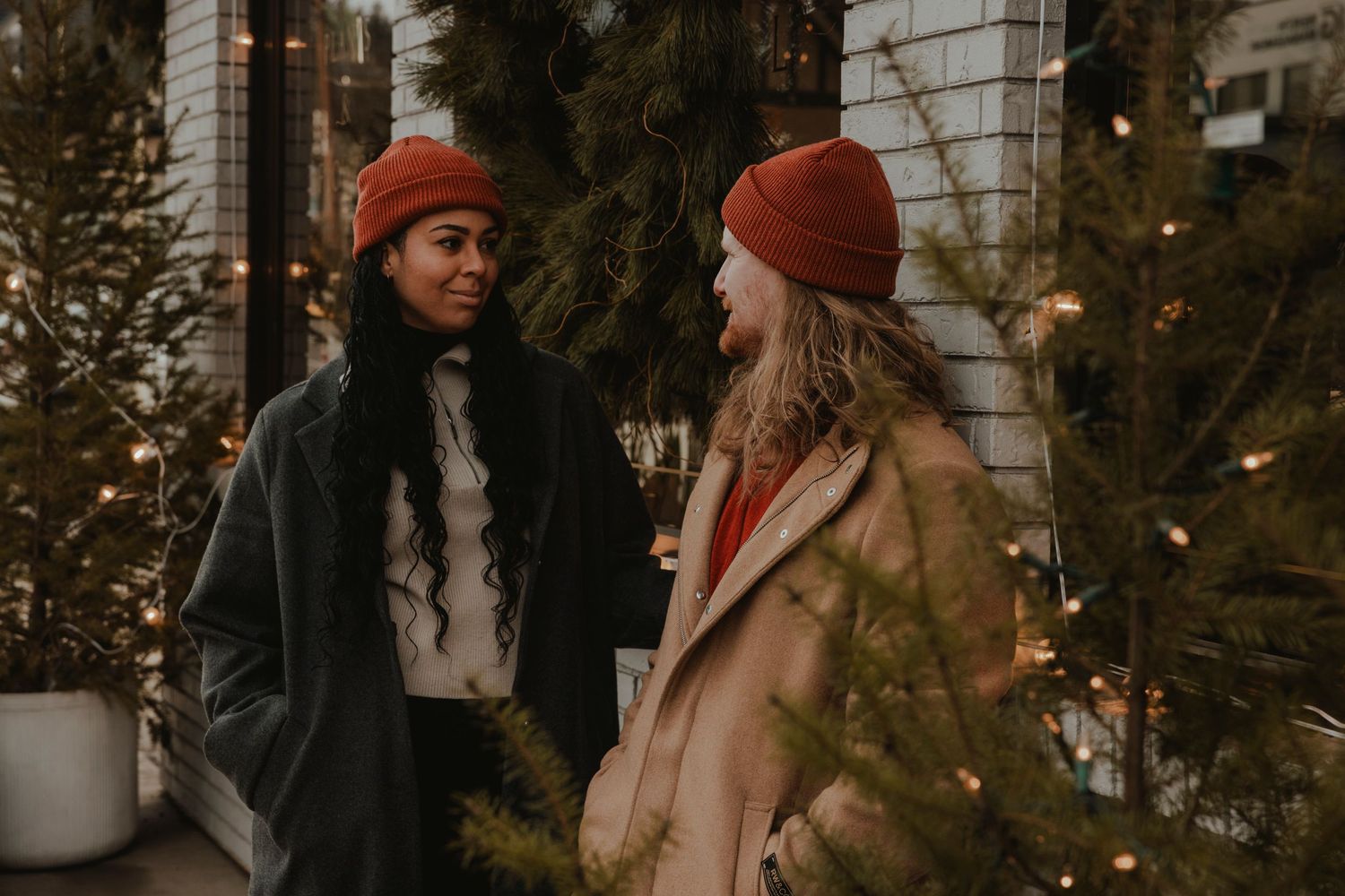 Winter couple photoshoot with red beanies against festive holiday decorations.