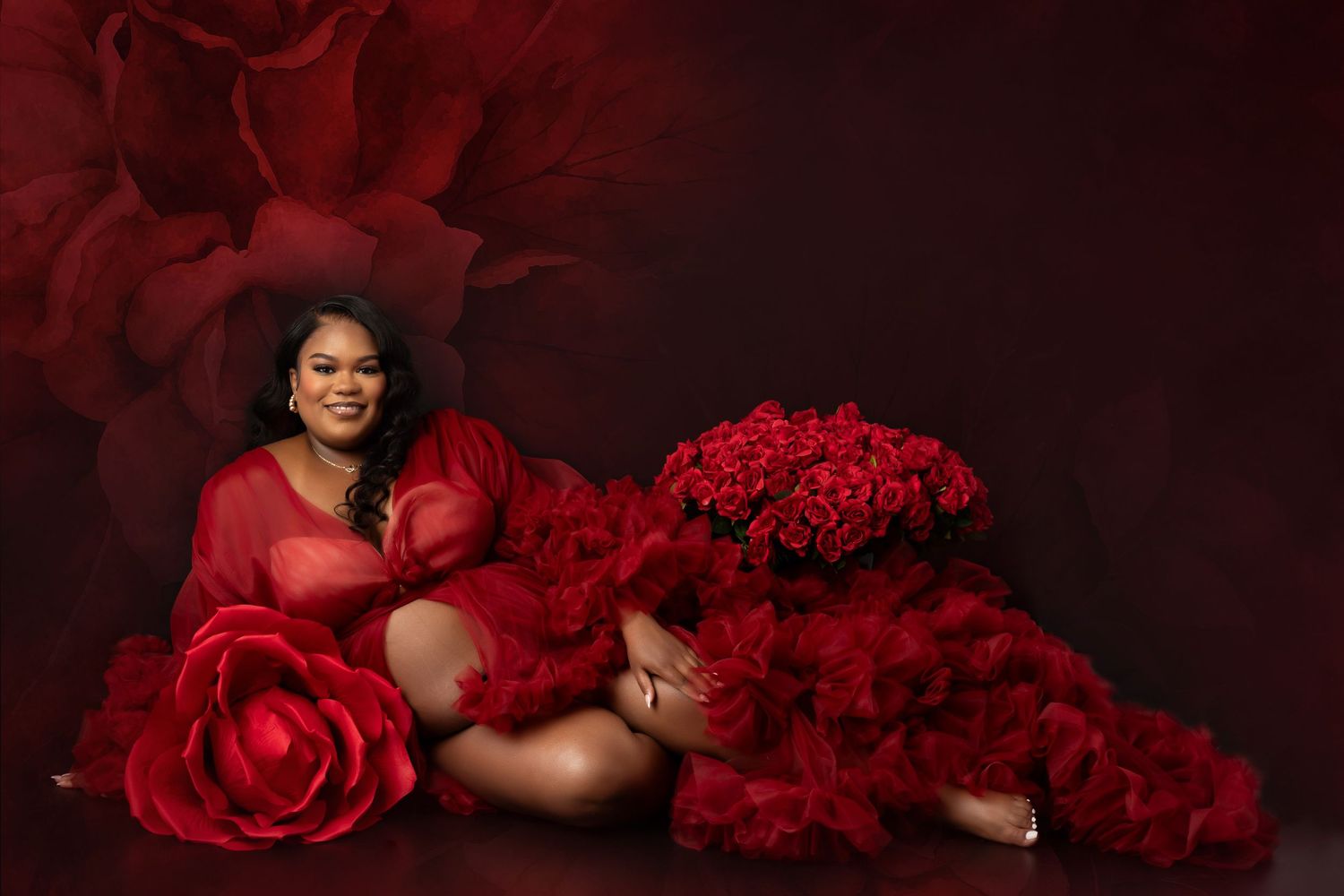 Woman in red ruffled gown poses elegantly among red roses and floral backdrop in a stunning maternity photo shoot.