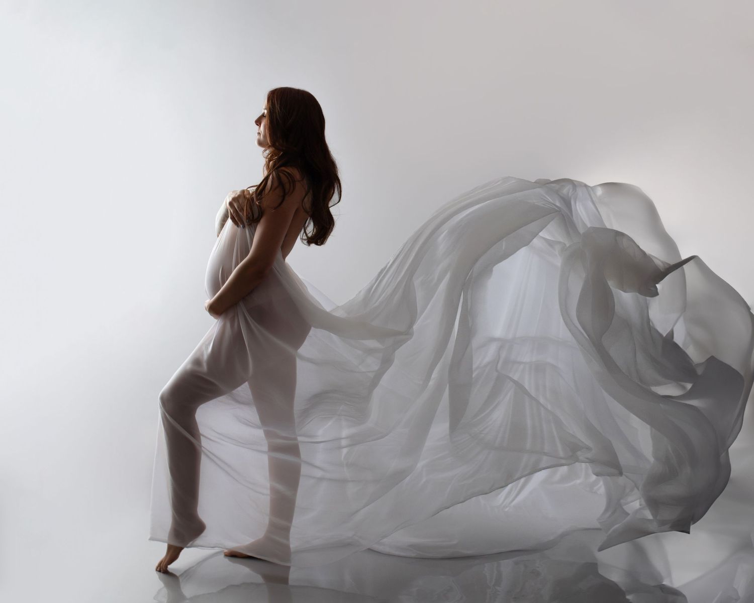 Pregnant woman in flowing white sheer fabric dress poses gracefully against a white studio background.