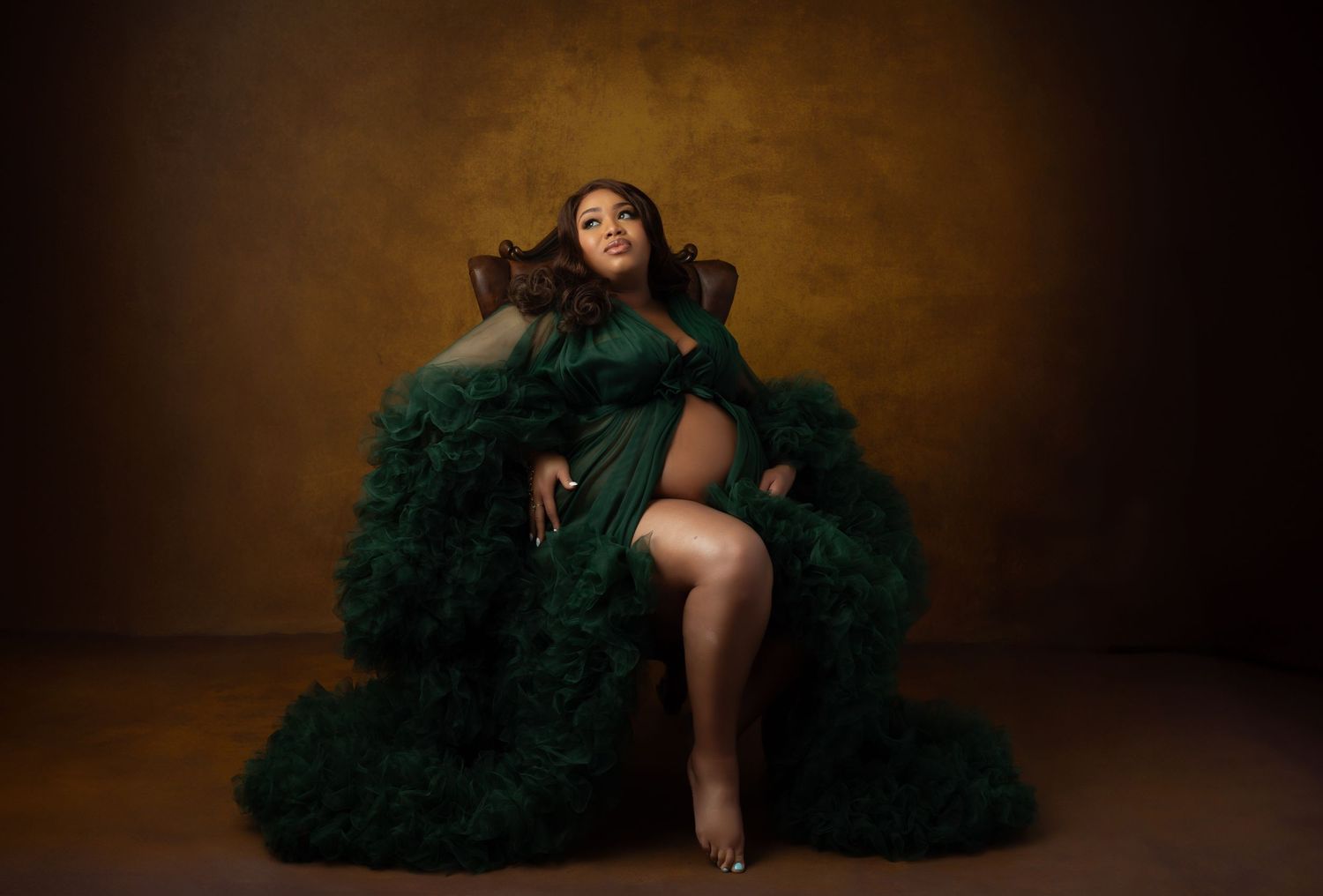 Pregnant woman in elegant dark green tulle gown seated in chair against warm brown studio backdrop.