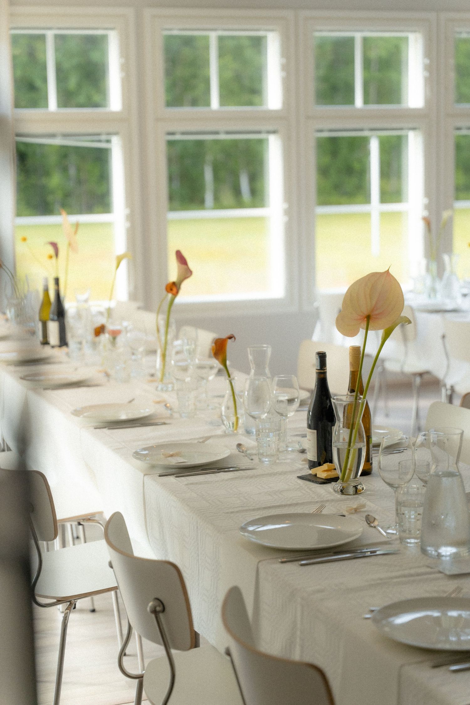 Elegant white dining table set with plates, wine glasses and calla lily centerpieces in a bright sunlit room with large windows.