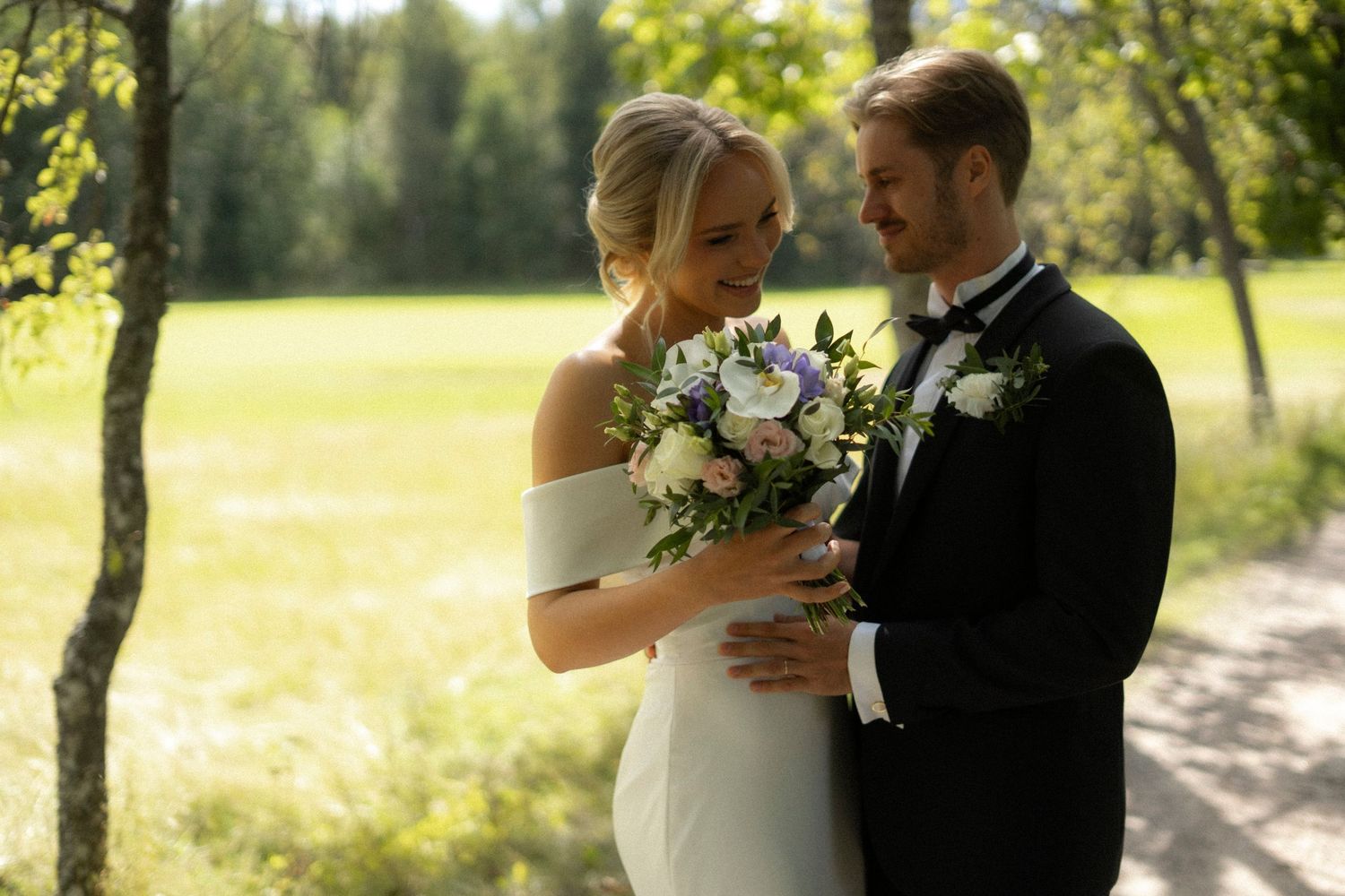 A couple in formal wedding attire embraces lovingly in a sunlit outdoor setting with trees and grass in the background.