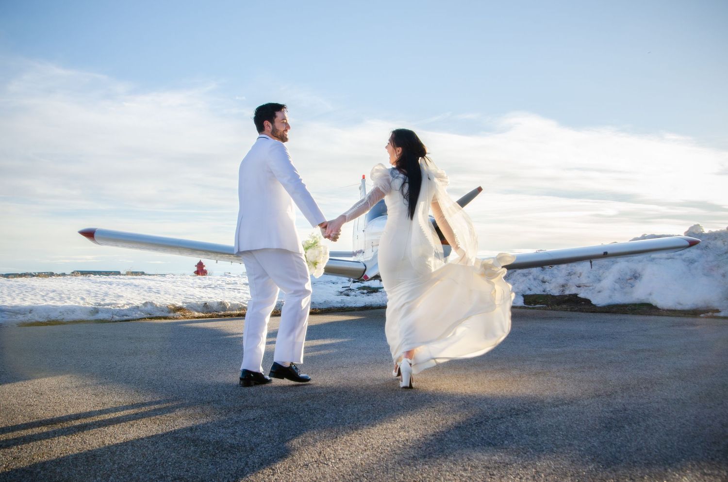 A couple in white wedding runs towards a small plane during their elopement photoshoot at sunset.